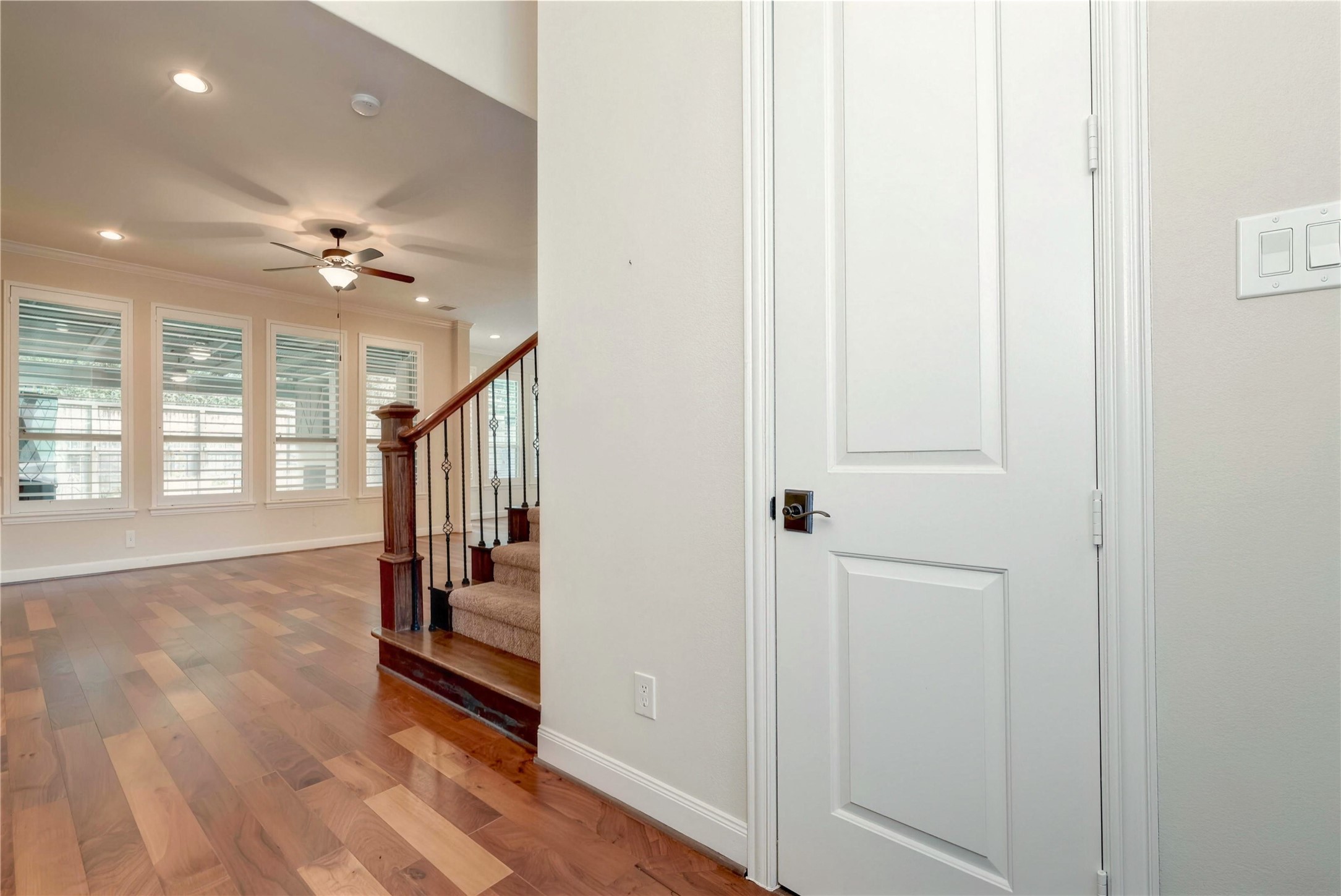 Elegant hardwood floors lead into a spacious living area, highlighted by large windows and a stylish ceiling fan, with an inviting staircase featuring wrought iron details.