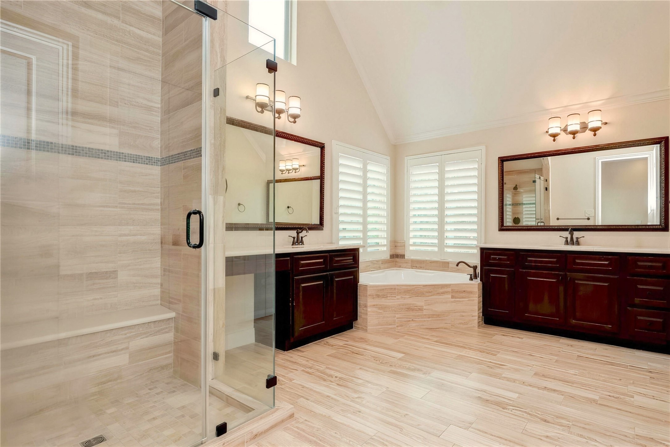 Elegant bathroom featuring a spacious glass-enclosed shower, luxurious soaking tub, dual vanities with rich wood cabinetry, and abundant natural light through plantation shutters.