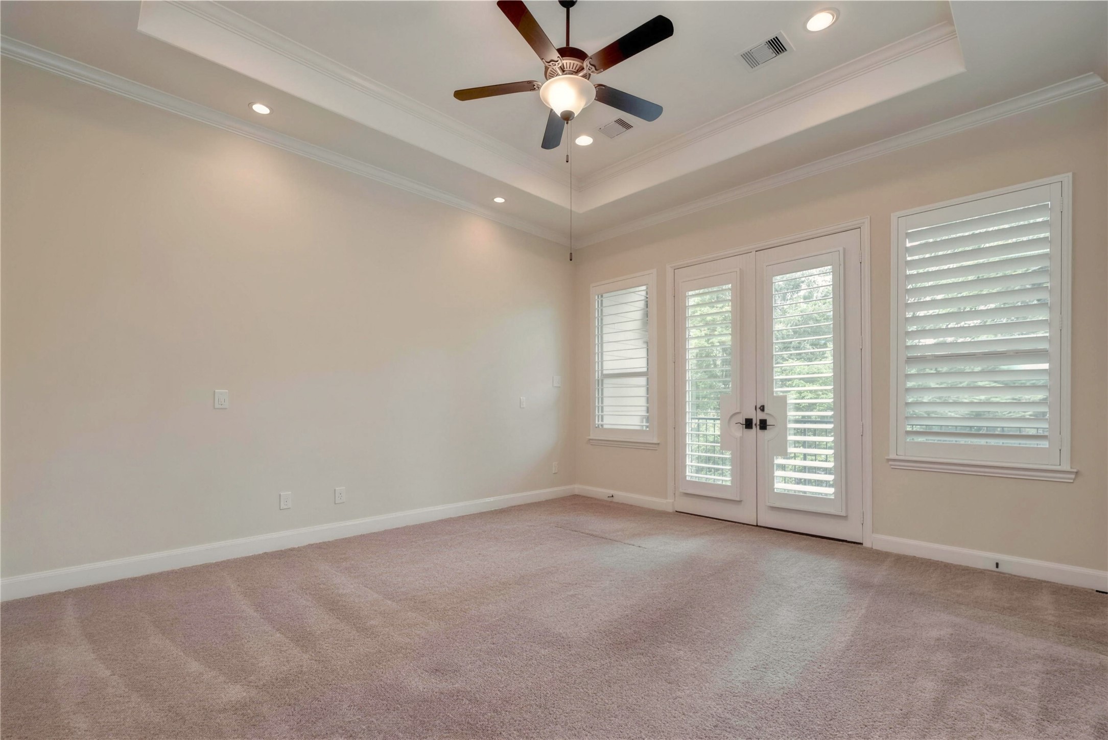 Spacious bedroom with a tray ceiling, elegant fan, and ample natural light streaming through French doors and large windows with shutters.