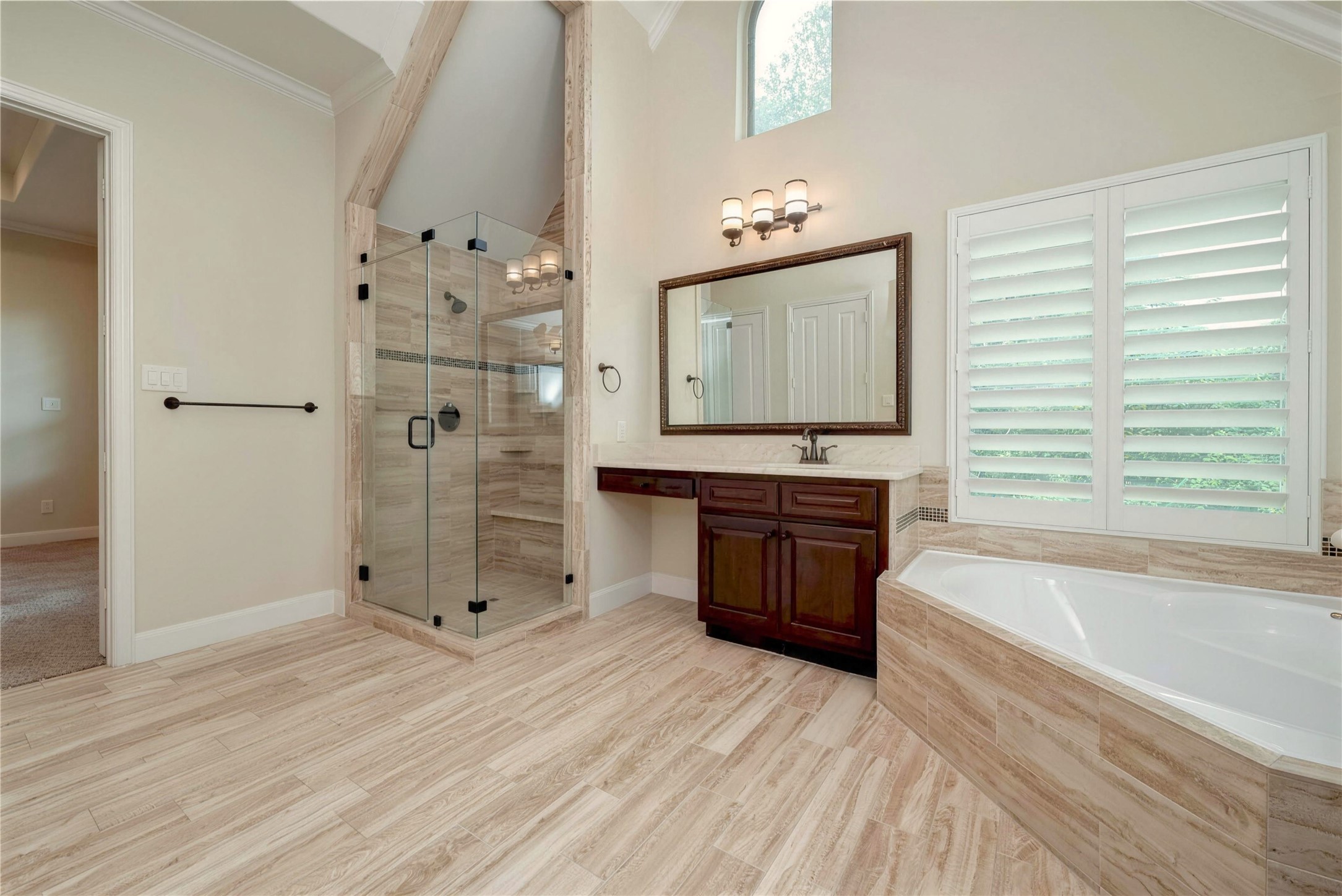Elegant bathroom featuring a spacious glass-enclosed shower, luxurious soaking tub, and rich wood vanity. Natural light pours in through large windows with plantation shutters, highlighting the serene atmosphere.