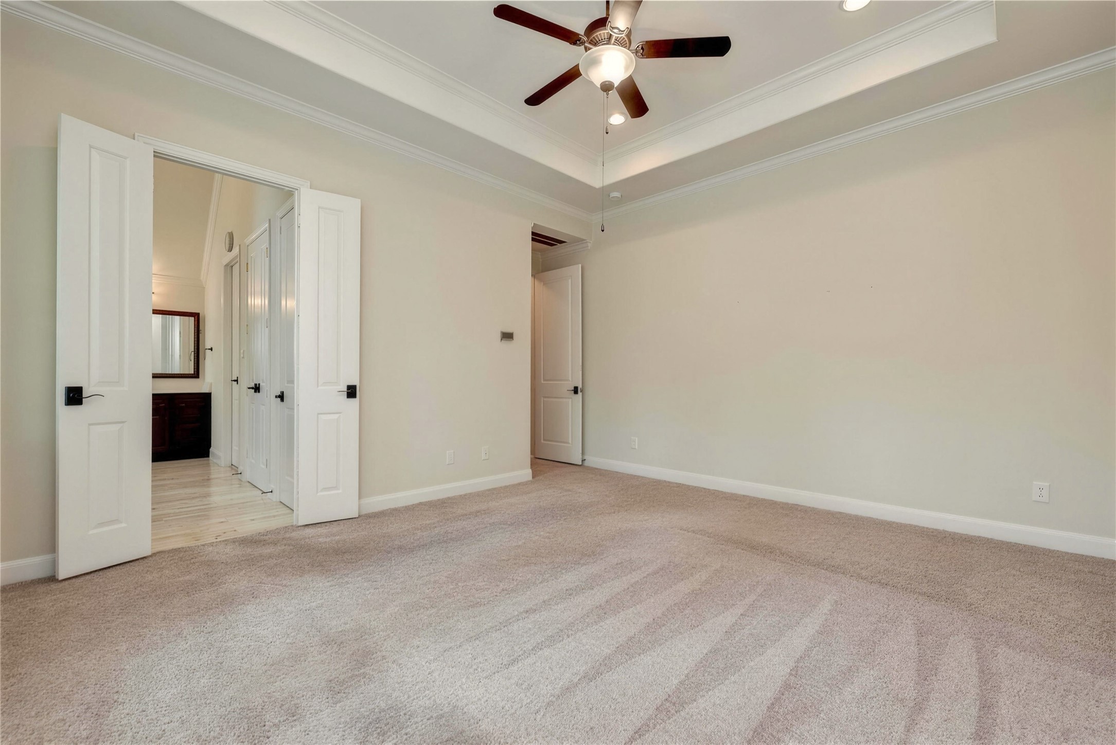 Spacious bedroom featuring plush carpeting, elegant tray ceiling with recessed lighting, and a stylish ceiling fan. Double doors open to an adjoining bathroom, adding convenience and luxury.