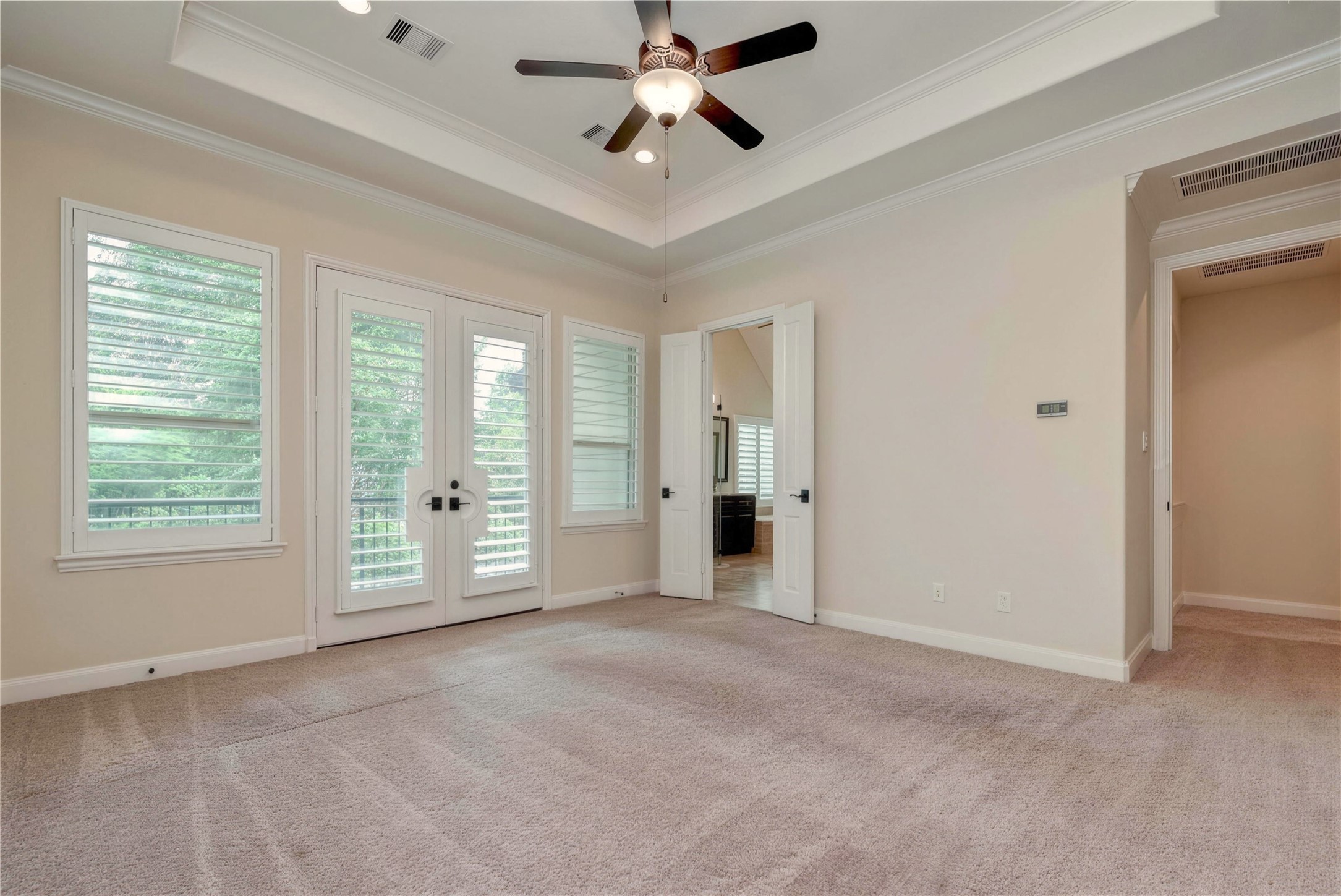 Spacious bedroom with a tray ceiling, elegant ceiling fan, and abundant natural light streaming through large windows and French doors, offering a serene view of the outdoors.
