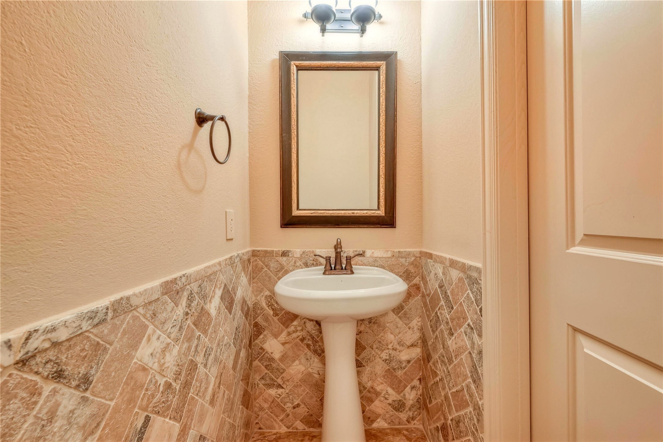Charming powder room featuring elegant stone tile walls, a classic pedestal sink, and a stylish framed mirror under soft lighting.