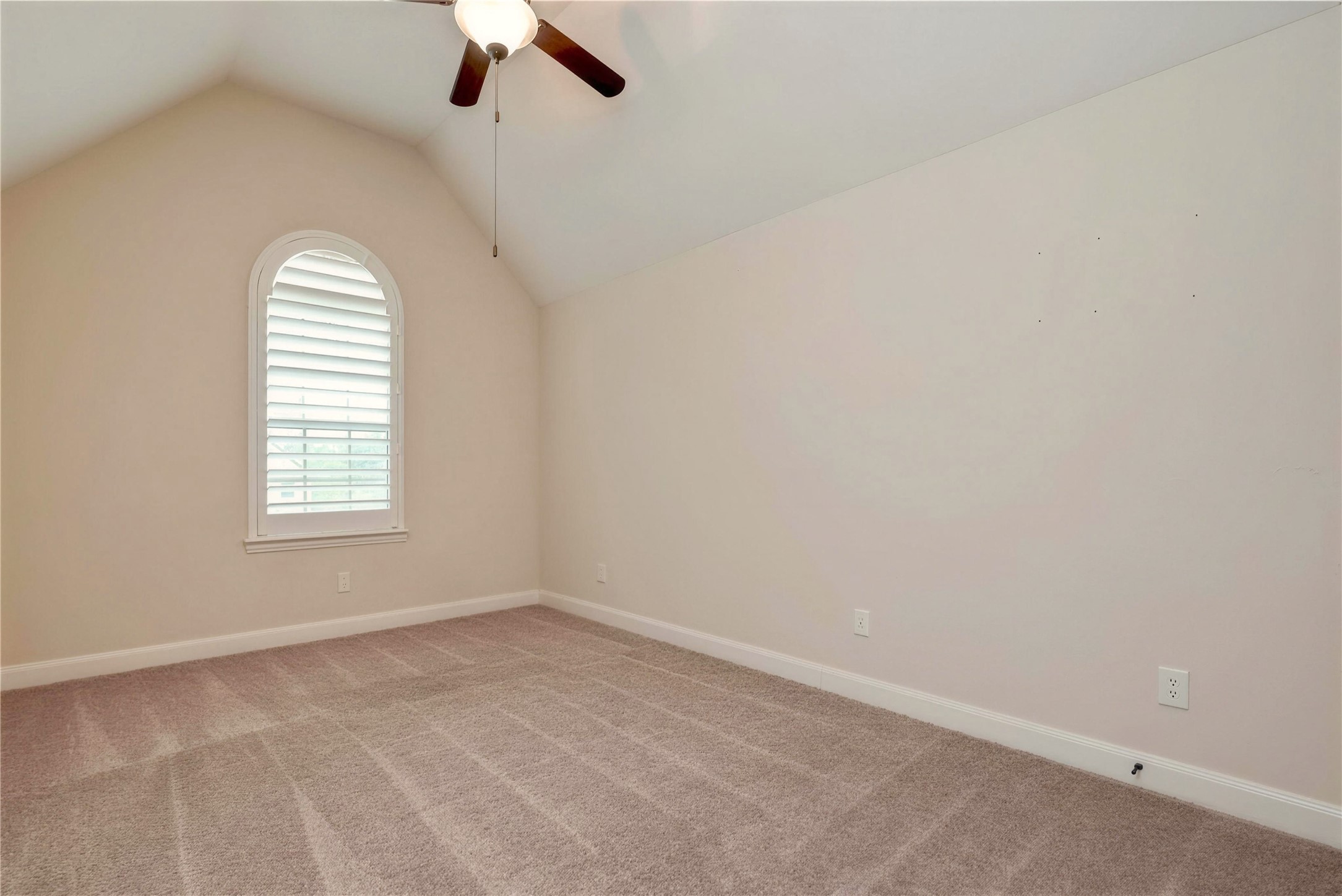 Bright, airy room with vaulted ceiling, plush carpeting, and a charming arched window featuring plantation shutters.