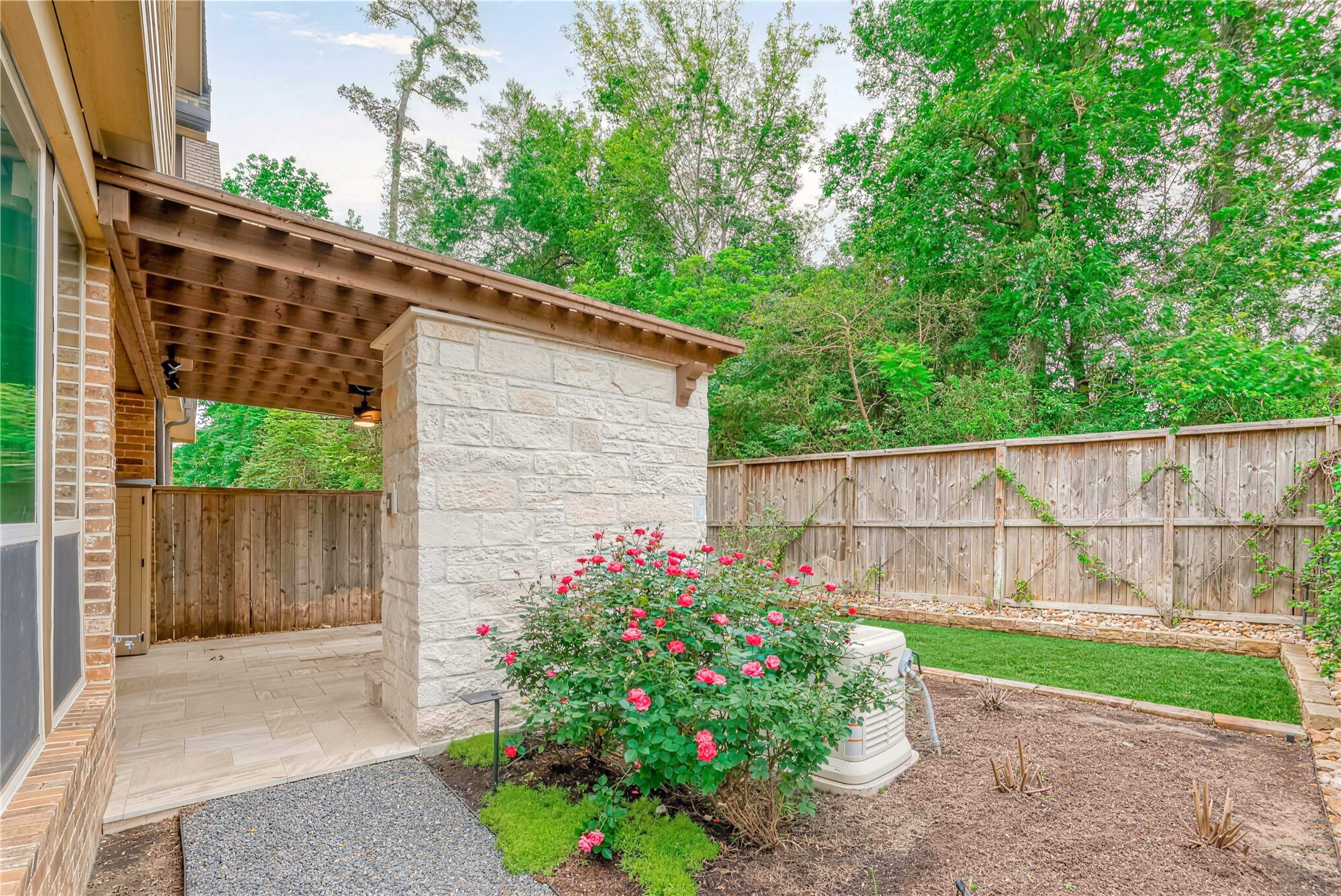 Charming outdoor space featuring a covered patio with elegant stonework, a lush garden with blooming roses, and a private fenced yard surrounded by mature trees.