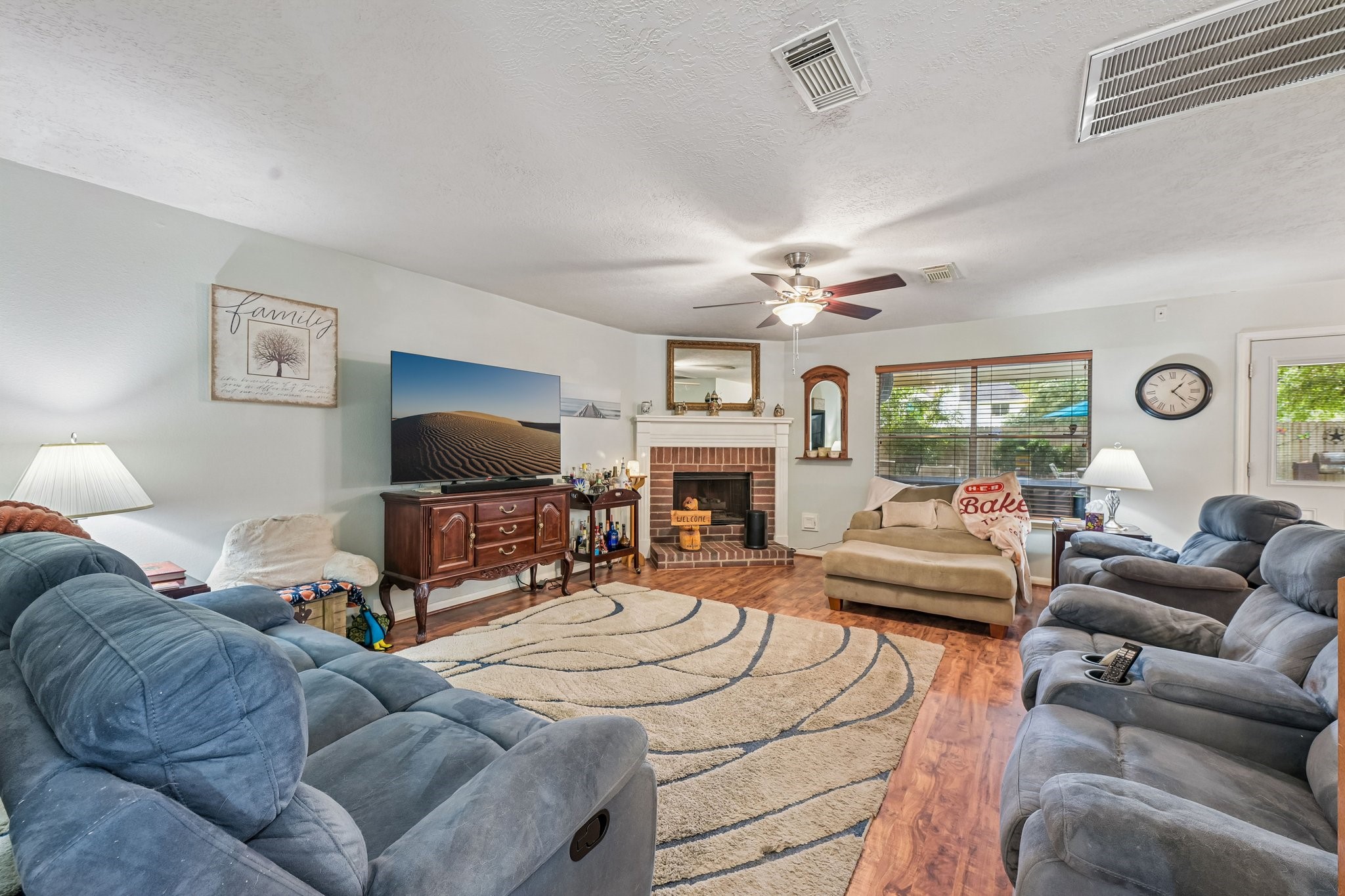 Expansive living room with wood-look flooring, ceiling fan, natural lighting, and a focal-point brick fireplace. Expansive living room with wood-look flooring, ceiling fan, natural lighting, and a focal-point brick fireplace.
