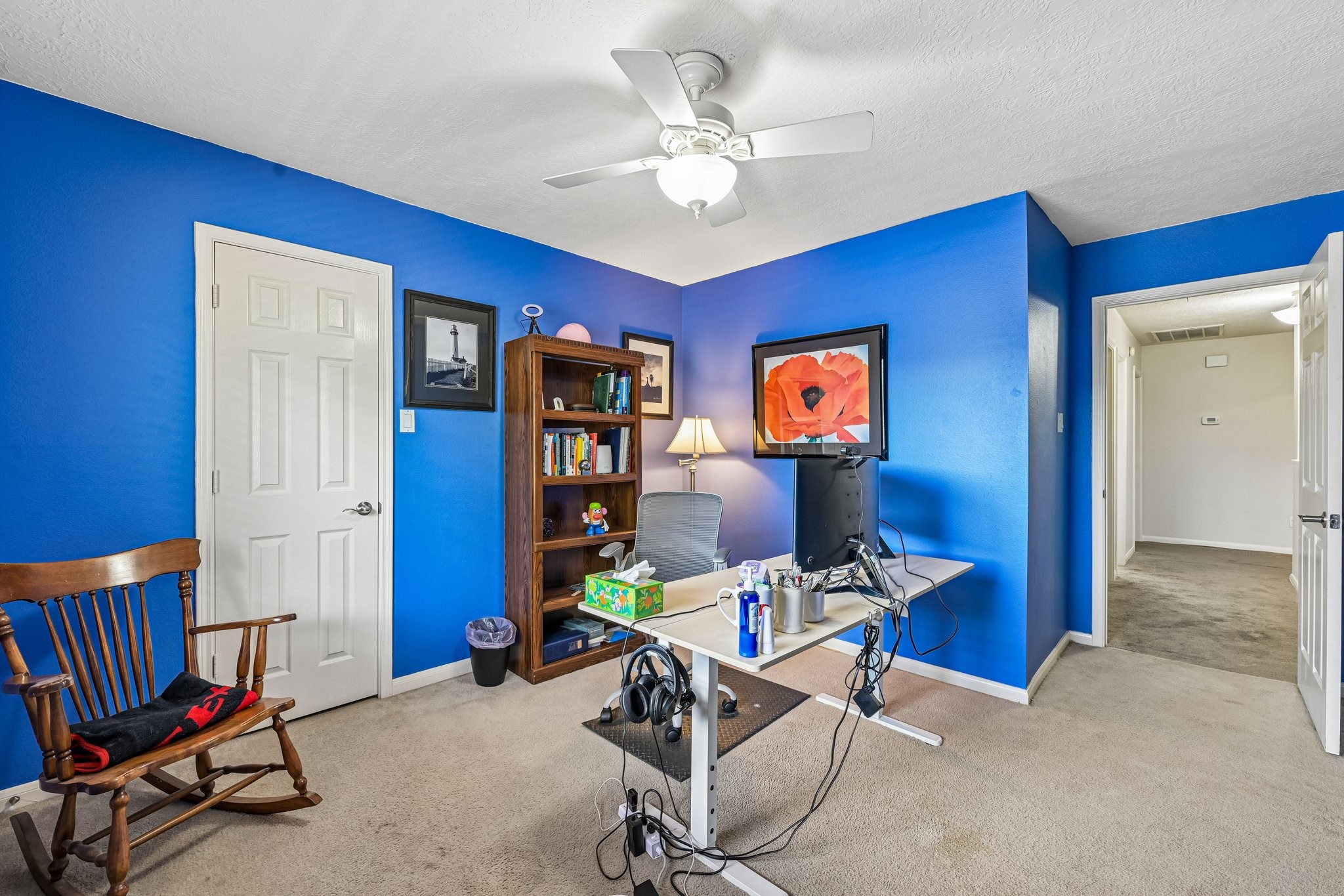 Spacious secondary bedroom/home office featuring vibrant blue accent walls, soft neutral carpeting, a ceiling fan, and direct hallway access.