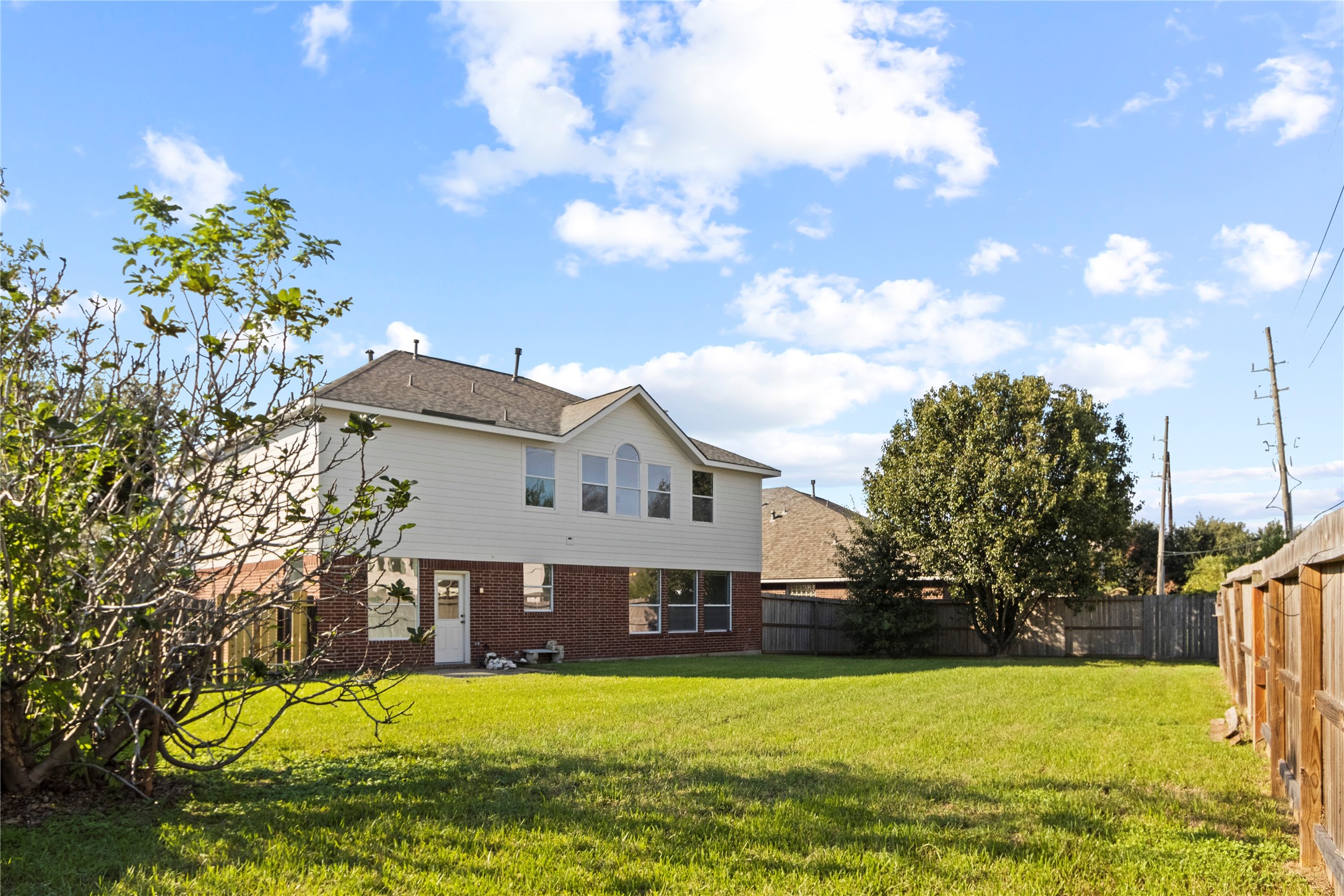 Spacious fenced backyard with mature trees and a privacy fence behind this two-story brick-and-siding home, featuring a wall of upstairs windows that flood the interior with natural light.