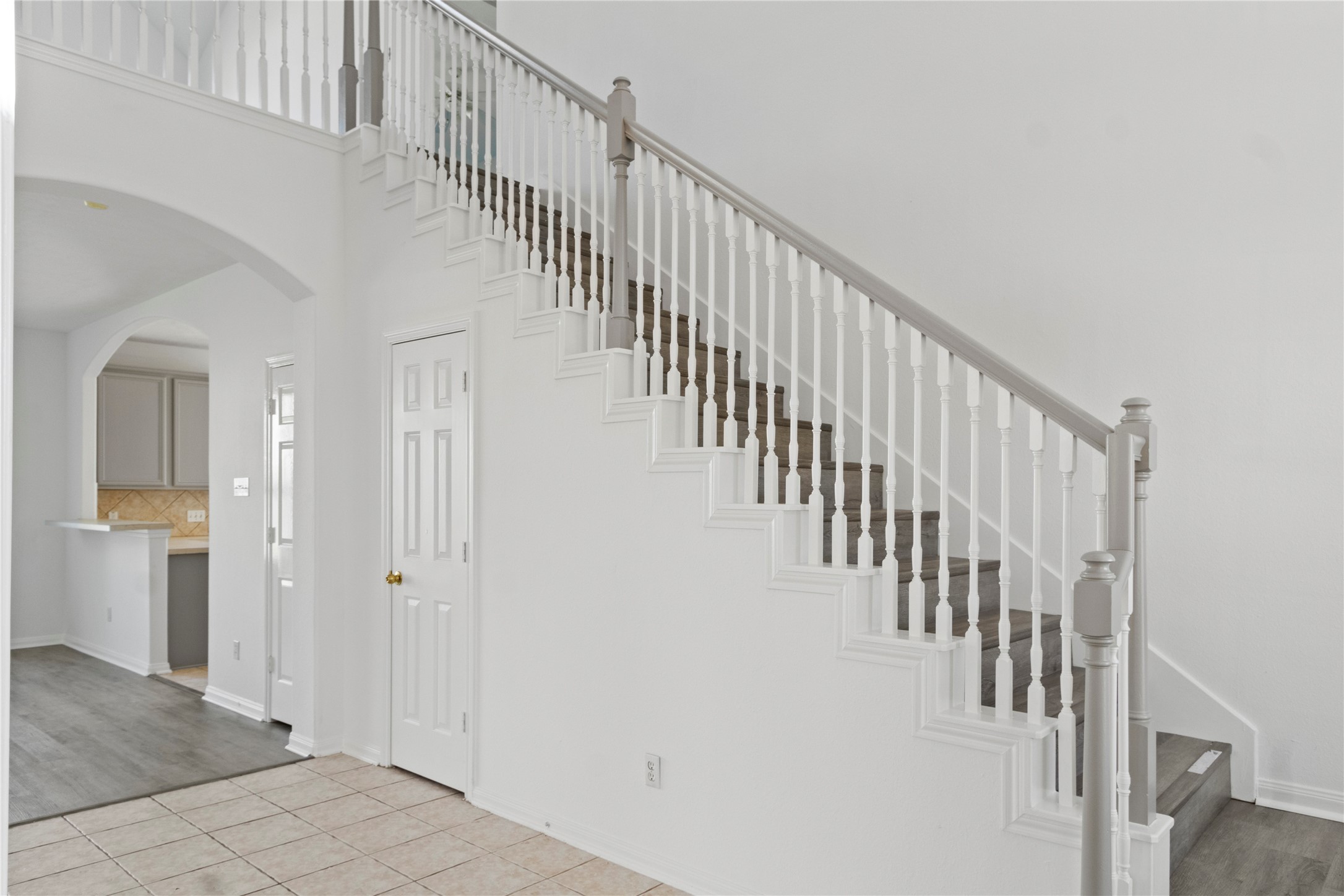 Interior view showcasing a staircase with white balusters and dark-toned steps, complemented by decorative trim and neutral walls. Tile flooring in the entryway transitions to gray-toned flooring in the adjoining living area, with an arched opening leading toward the kitchen.