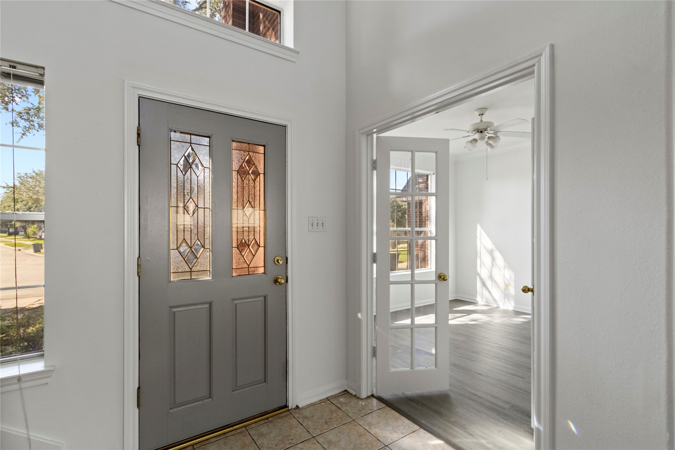 Front entry featuring a decorative glass front door, tile flooring, and large windows providing natural light. An adjacent room with gray-toned flooring, a ceiling fan, and French doors offers a versatile space for a home office, formal dining room, or additional living area.