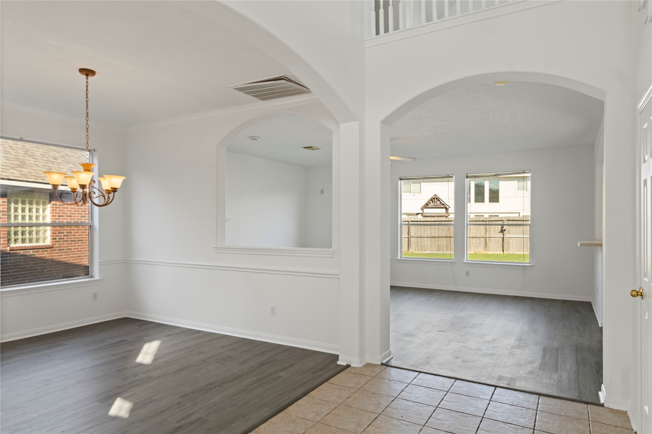 Open-concept living and dining areas featuring arched entryways, an upper-level railing, neutral interior finishes, and gray wood-look flooring. Large windows provide natural light, while the adjoining spaces offer a functional, connected layout. Tile flooring at the entry adds a distinct transition between areas.