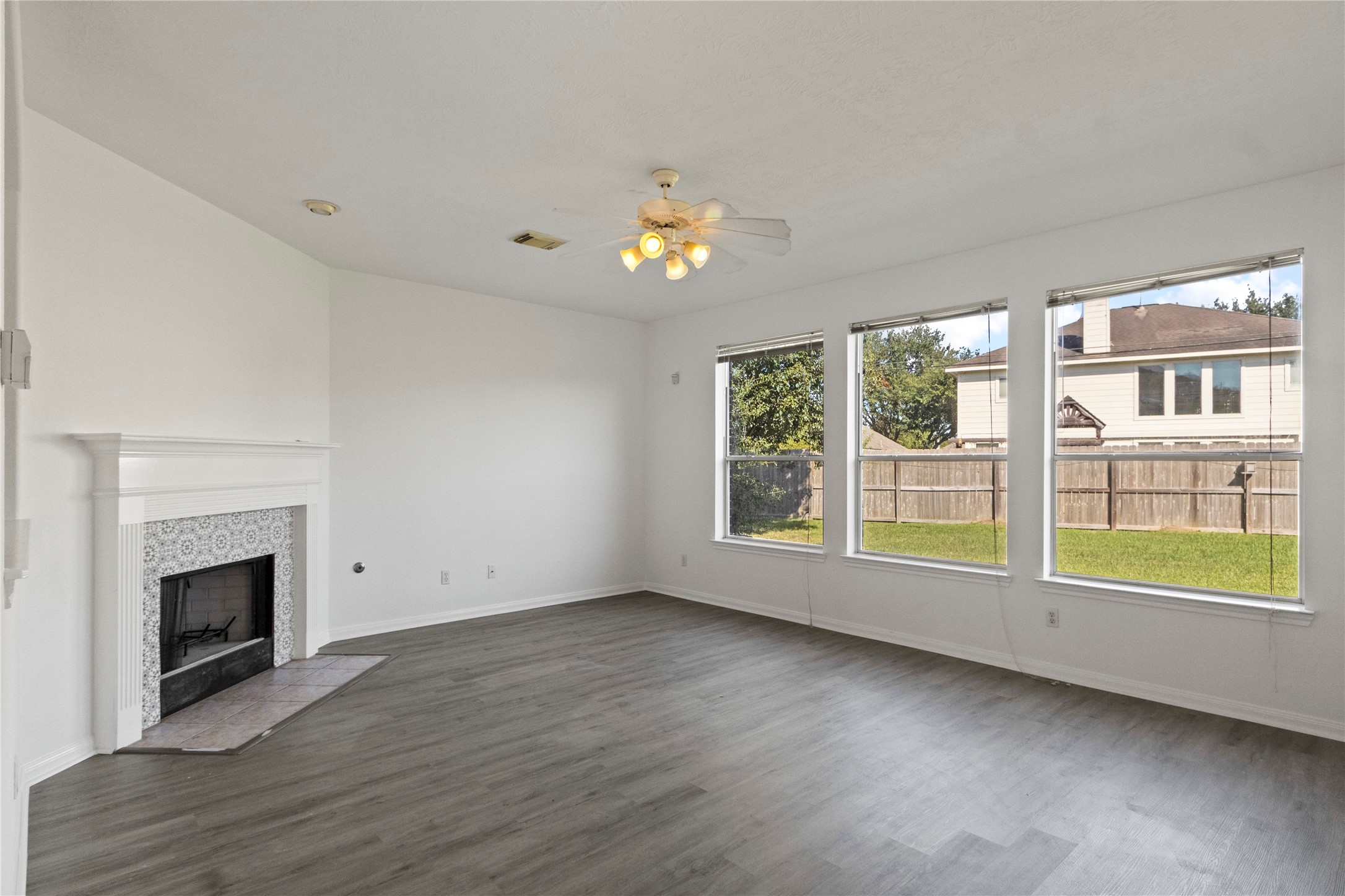 Spacious living room featuring a fireplace with decorative tile surround, gray wood-look flooring, and a ceiling fan with light fixture. Three large windows provide abundant natural light and views of the backyard. Neutral walls and an open layout offer a versatile space for relaxing and entertaining.
