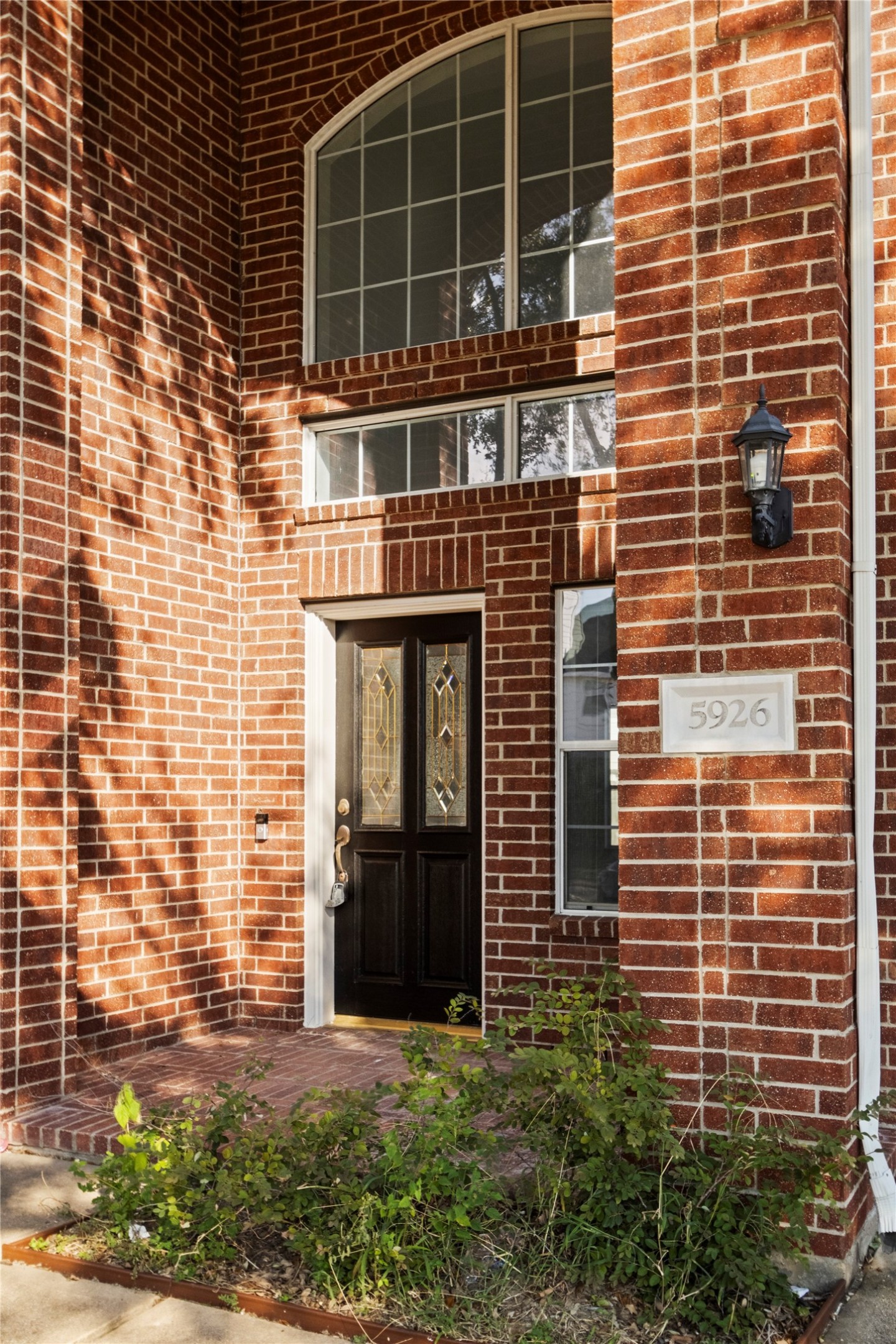 Covered front entry featuring a decorative glass front door, an arched upper window, and red brick detailing. A brick-paved porch and surrounding landscaping complete the welcoming entrance.