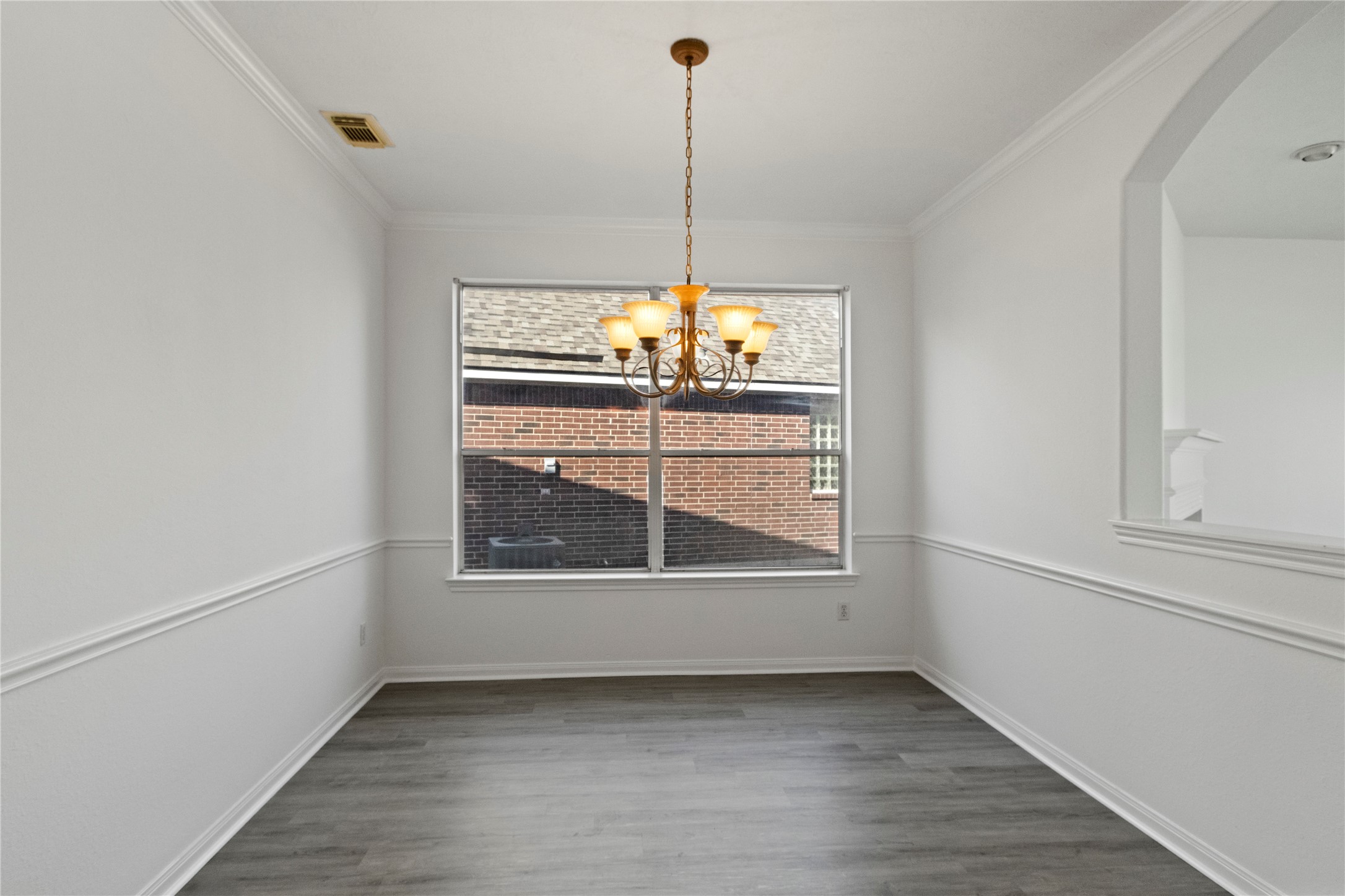 Formal dining room featuring a chandelier, crown molding, chair rail detailing, neutral walls, and gray wood-look flooring. Large window provides natural light, while the open pass-through offers a seamless connection to the adjacent living area.
