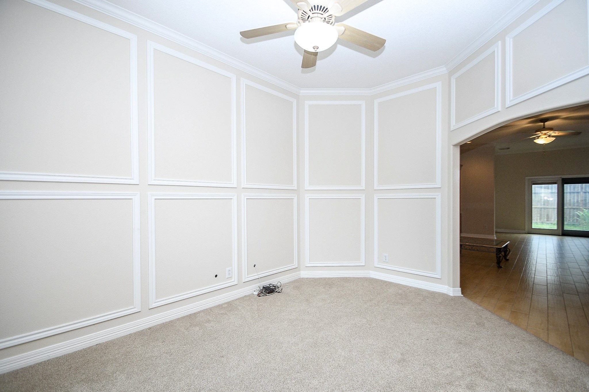 This room features neutral-toned walls with elegant paneling, a plush carpet, and a ceiling fan. It opens up to a larger space with hardwood floors and a view of the backyard through large windows, creating a bright and welcoming atmosphere.