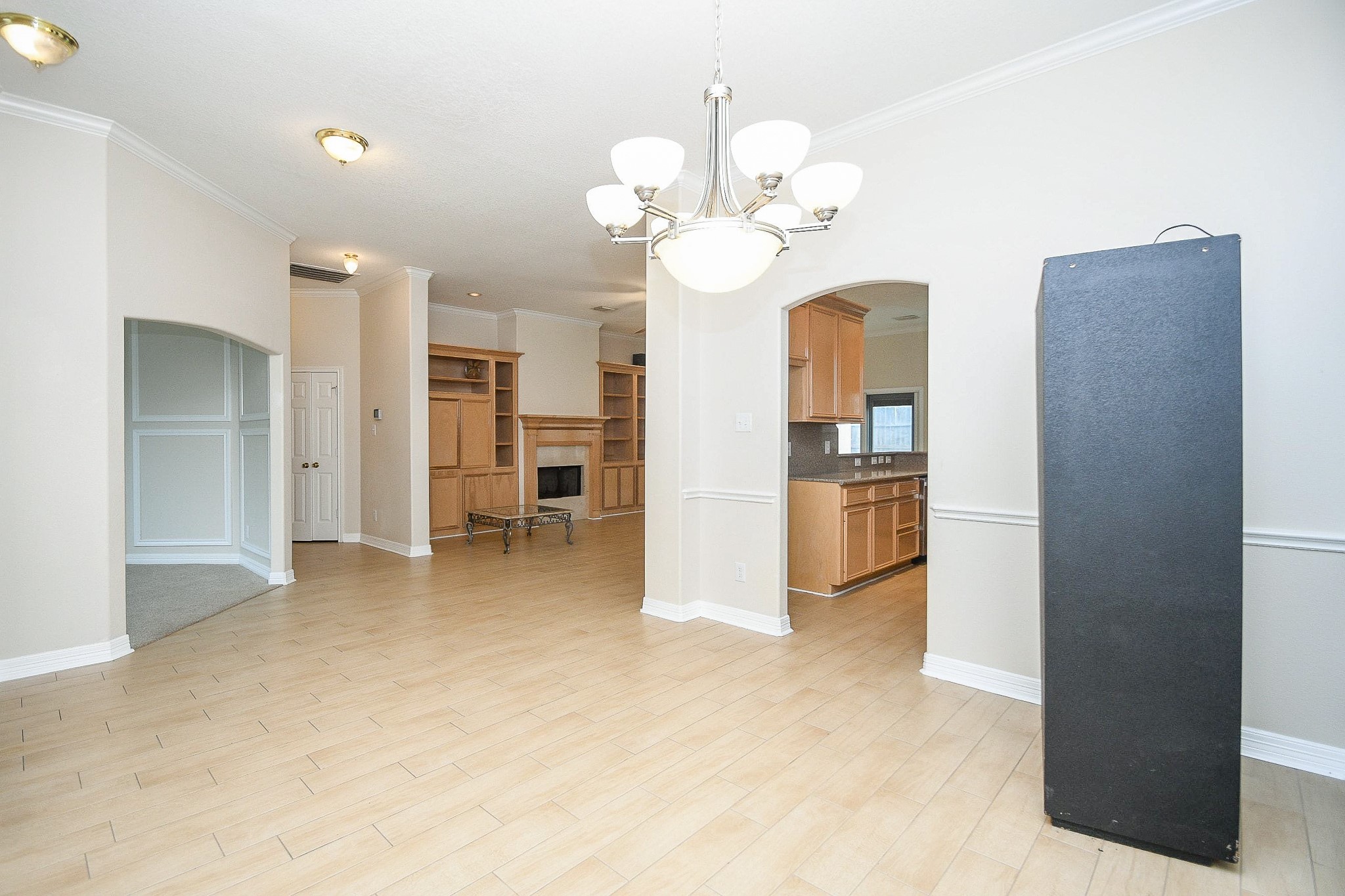 This spacious living area features light wood flooring, neutral walls, a built-in fireplace, and elegant lighting fixtures. It seamlessly connects to the kitchen through an arched doorway, creating a welcoming and open atmosphere.
