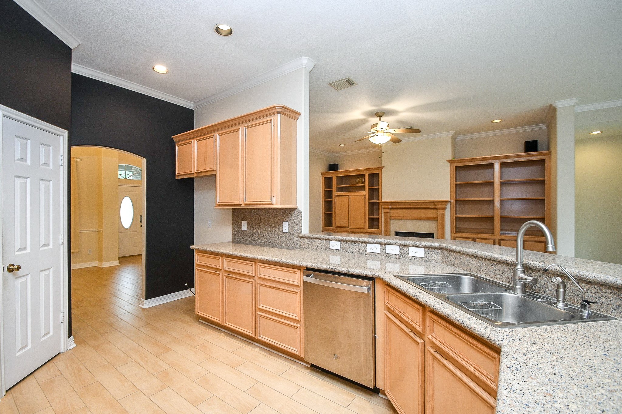 Bright and spacious kitchen with light wood cabinets, granite countertops, and a double sink. An open layout leads to a living area with built-in shelving and a fireplace. Elegant archway and ample natural light enhance the welcoming atmosphere.