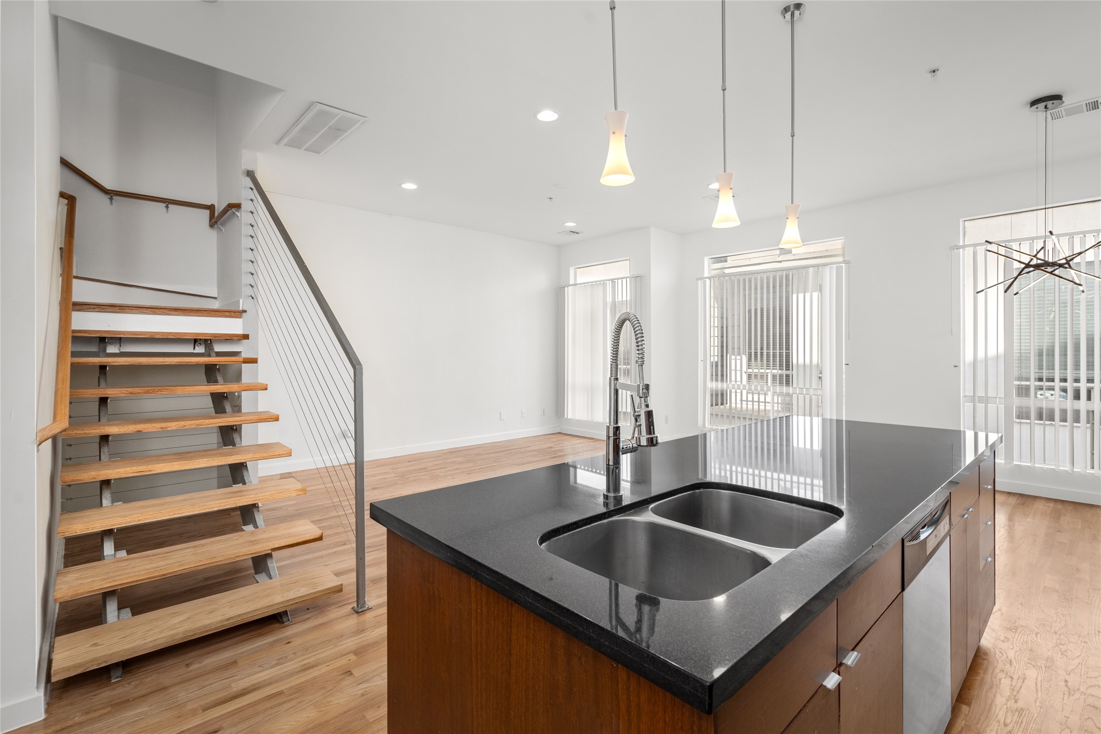 A light hardwood floor covers the space. A wooden staircase with metal railings leads up, its open design showing the support structure.

In the center, a modern kitchen island stands with a dark granite countertop, a stainless steel double sink, and a sleek faucet. The island has medium-brown wood cabinets, and a dishwasher is partly visible on the right.