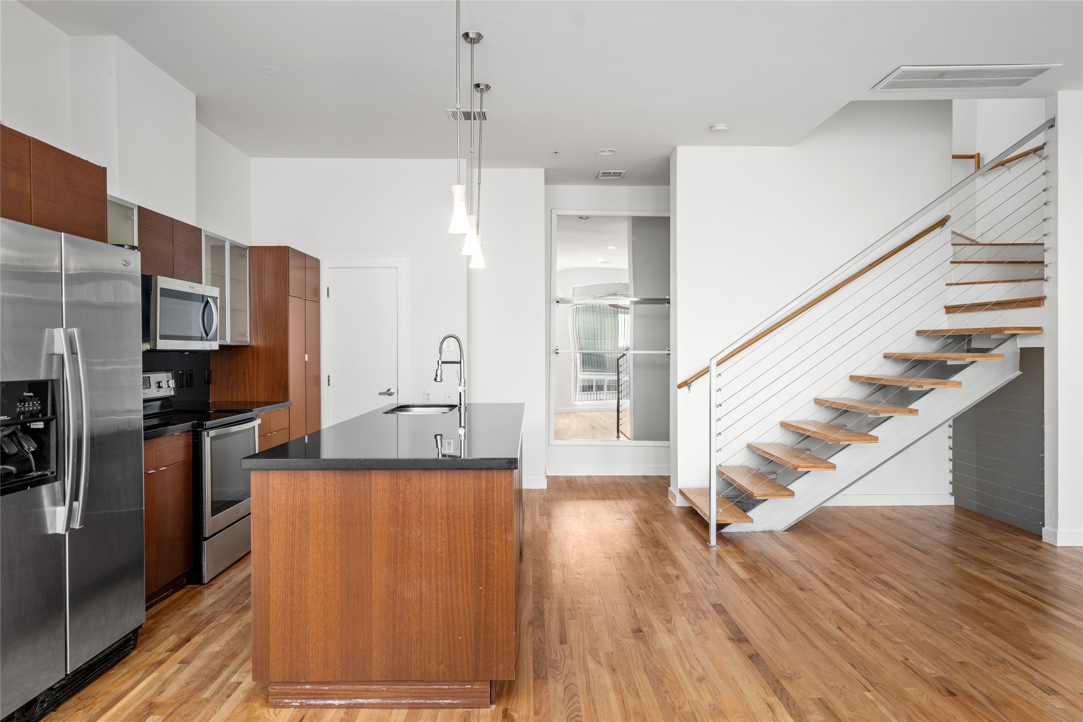 Stunning modern kitchen with sleek design and high-end finishes! Featuring rich dark-brown wood cabinetry, glossy black granite countertops, and stainless steel appliances, including a refrigerator, microwave, and oven. A spacious island with matching cabinetry provides extra prep space and seating.

Light hardwood floors flow seamlessly into the adjacent open staircase, which boasts white wooden steps, a stylish white metal railing, and thin wire-like supports. The neutral-toned walls and trio of pendant