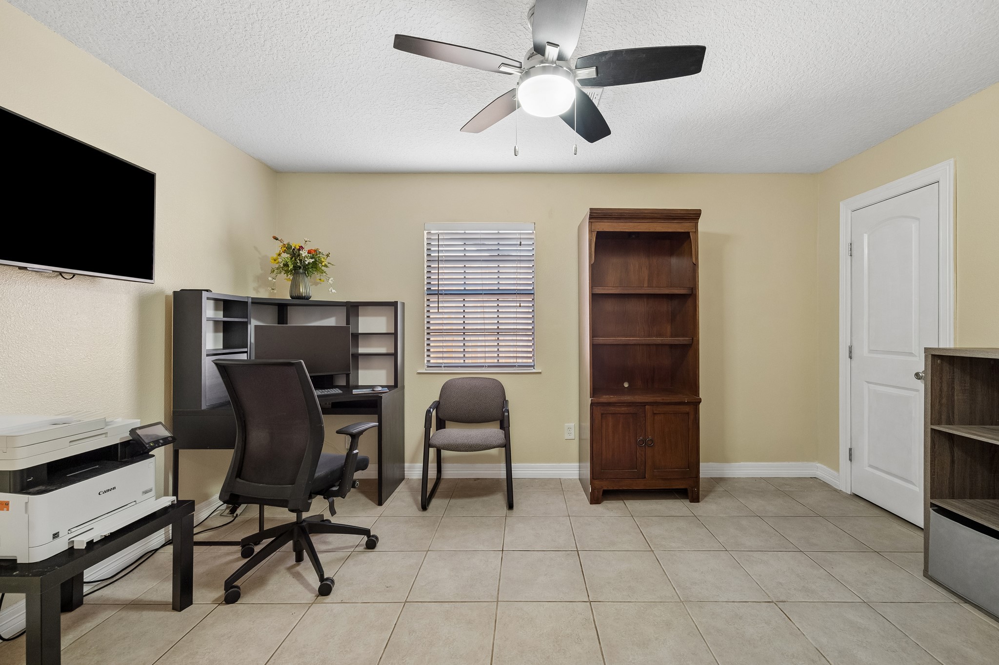 Bright and functional home office with neutral walls, tiled flooring, and ample storage space, featuring a ceiling fan for comfort.