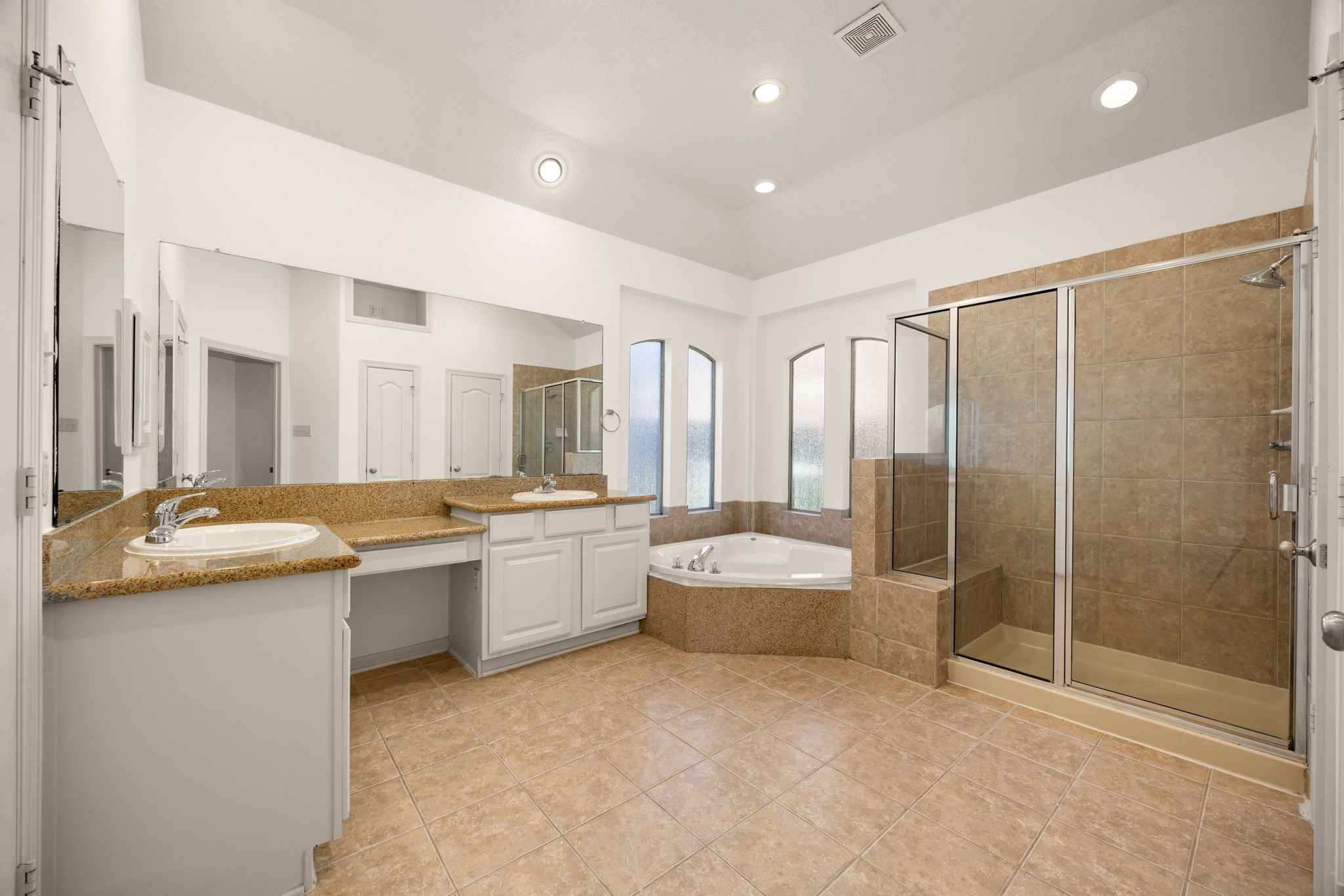 Designed as a spa-like retreat, this expansive primary bathroom features a generous double-vanity layout with a built-in makeup station, neutral granite countertops, clean white cabinetry, a deep soaking tub, and a large walk-in shower.