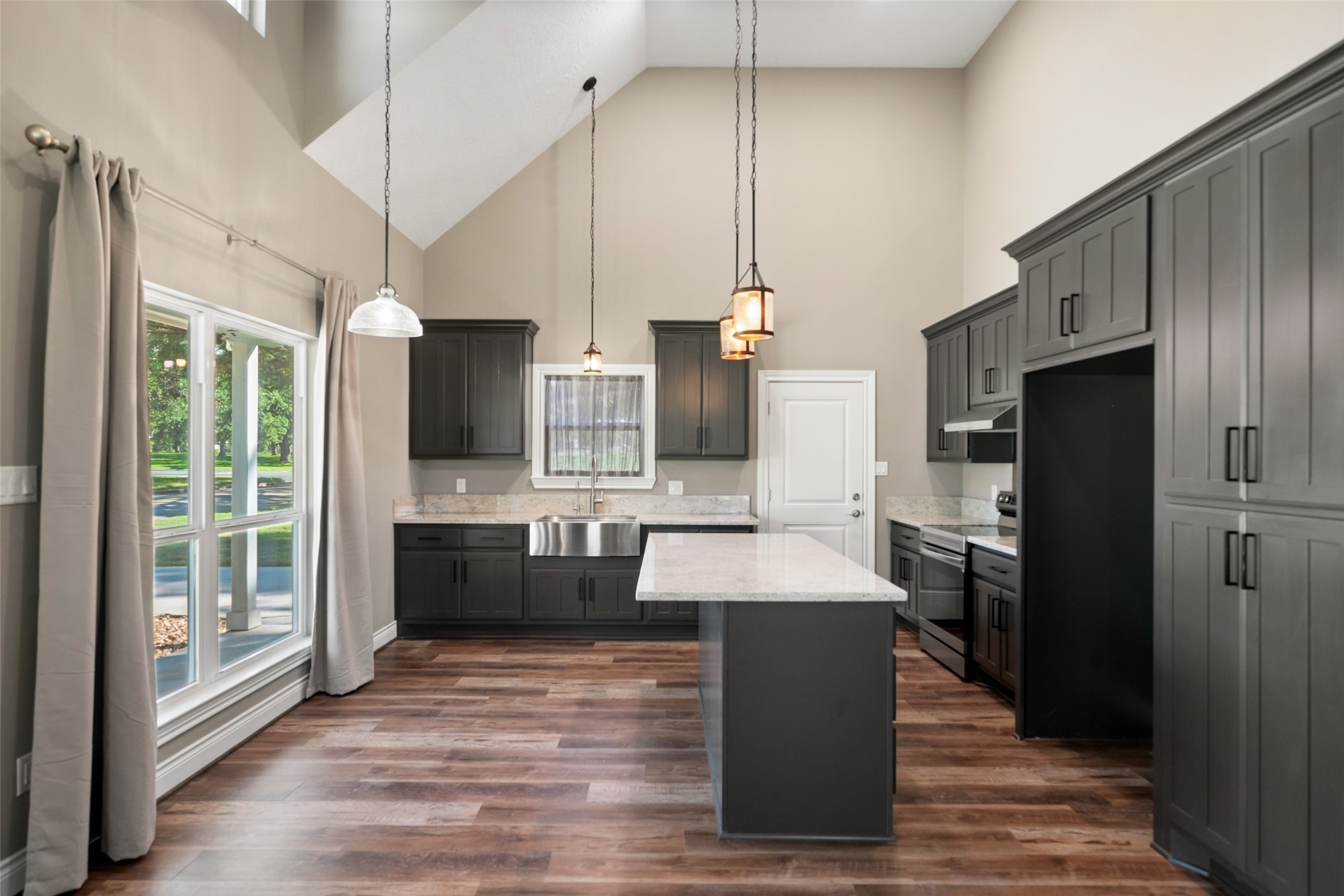 Kitchen featuring dark cabinetry, a center island, pendant lighting, and a stainless steel apron-front sink beneath the window.