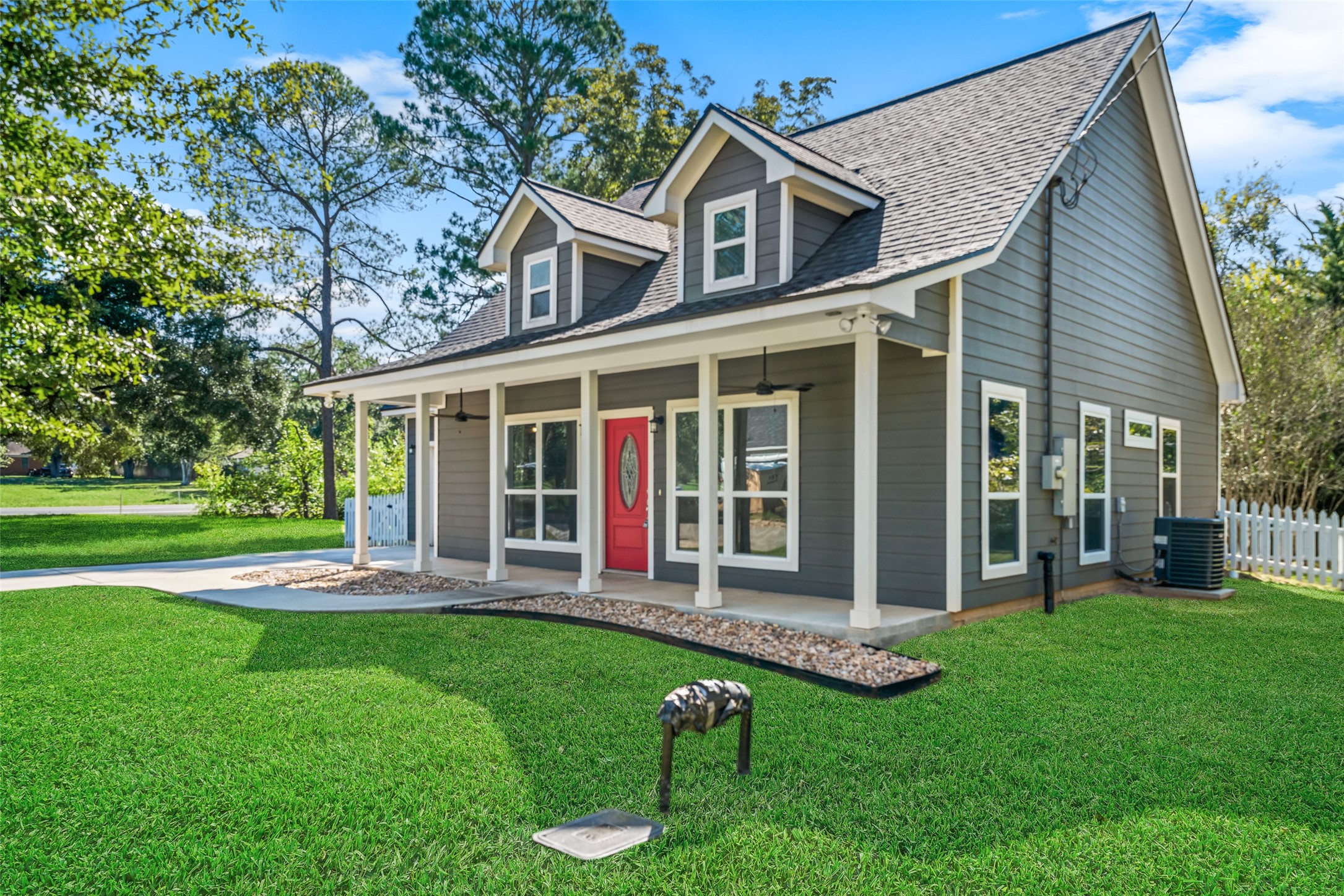Angled front view highlighting the covered porch, red entry door, dormer windows, and mature shade trees.