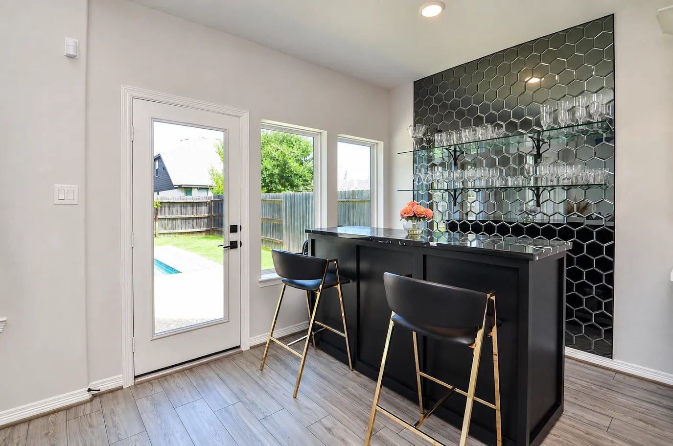 Stylish home bar with sleek black hexagonal backsplash, well-stocked glass shelves, and a chic counter overlooking the backyard pool area. Ideal for entertaining. Stylish home bar with sleek black hexagonal backsplash, well-stocked glass shelves, and a chic counter overlooking the backyard pool area. Ideal for entertaining.