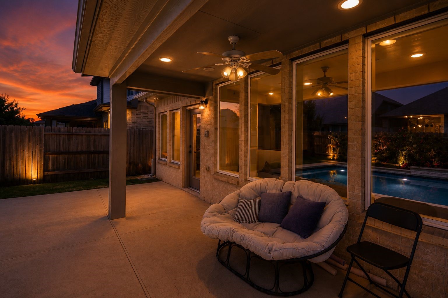 Experience serene evenings on the covered patio, featuring a cozy seating area and ceiling fan, overlooking a beautifully lit pool and sunset views.