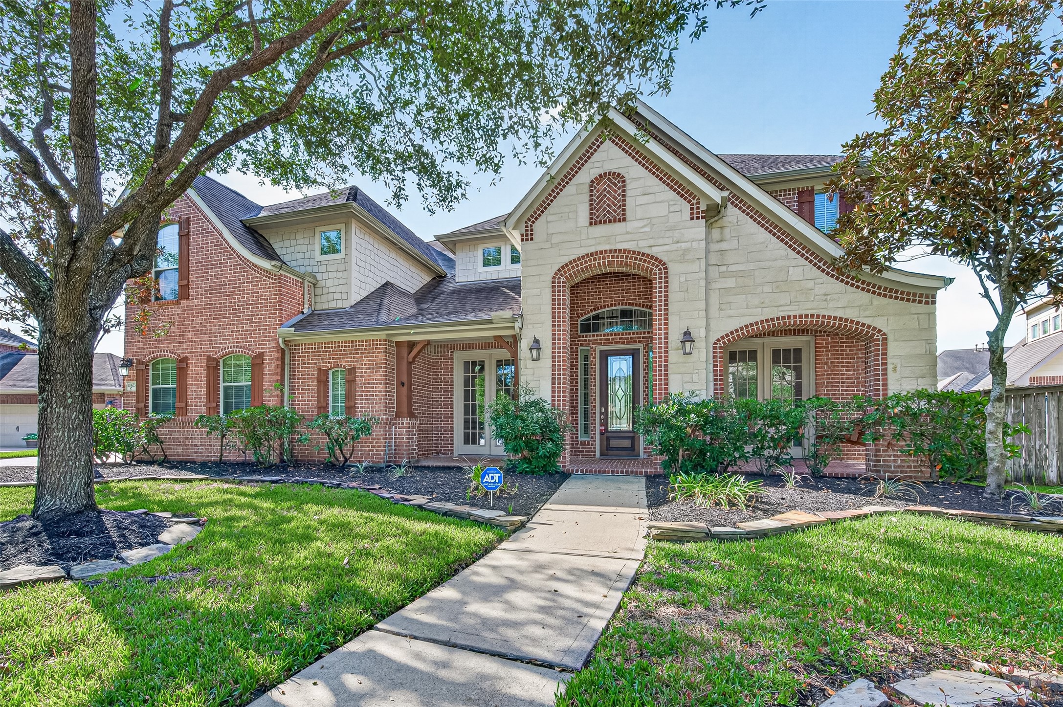 Charming two-story home with a blend of brick and stone exterior, featuring a welcoming arched entryway and lush landscaping.