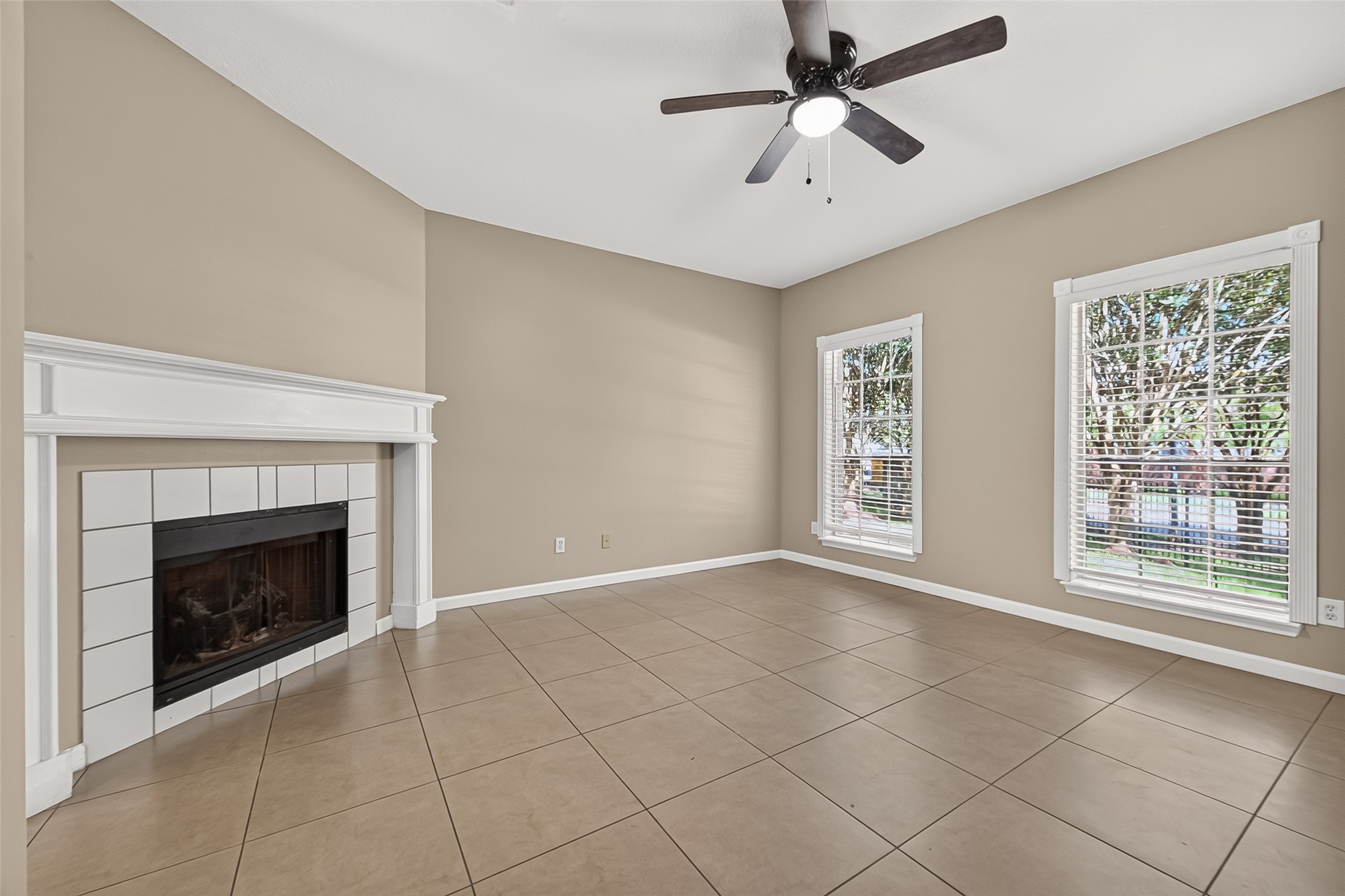 Inviting living room featuring a fireplace with decorative mantel, tile flooring, ceiling fan, and large windows overlooking the landscaped surroundings. Inviting living room featuring a fireplace with decorative mantel, tile flooring, ceiling fan, and large windows overlooking the landscaped surroundings.