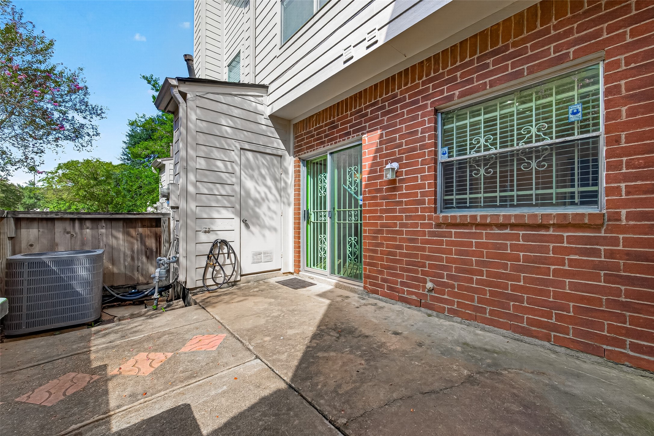 Fenced outdoor patio with brick exterior, mature shade trees, and convenient access to the home, offering a private space for relaxing or outdoor seating.