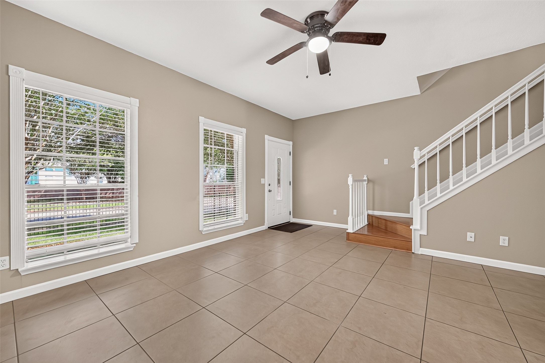 Spacious living area with tile flooring, ceiling fan, abundant natural light, and an attractive wood staircase with white railing. Spacious living area with tile flooring, ceiling fan, abundant natural light, and an attractive wood staircase with white railing.