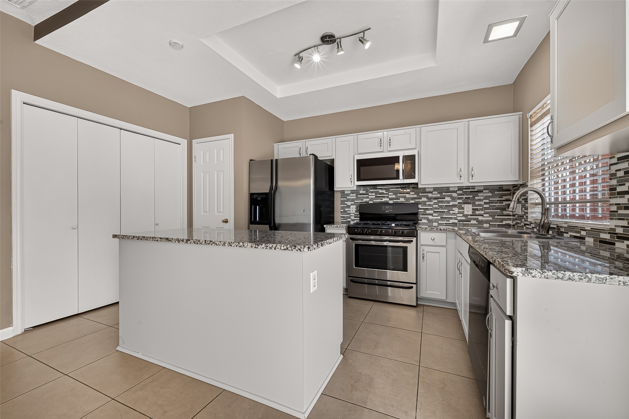 Bright kitchen with granite-topped island, stainless-steel appliances, white cabinetry, tile flooring, and contemporary lighting.