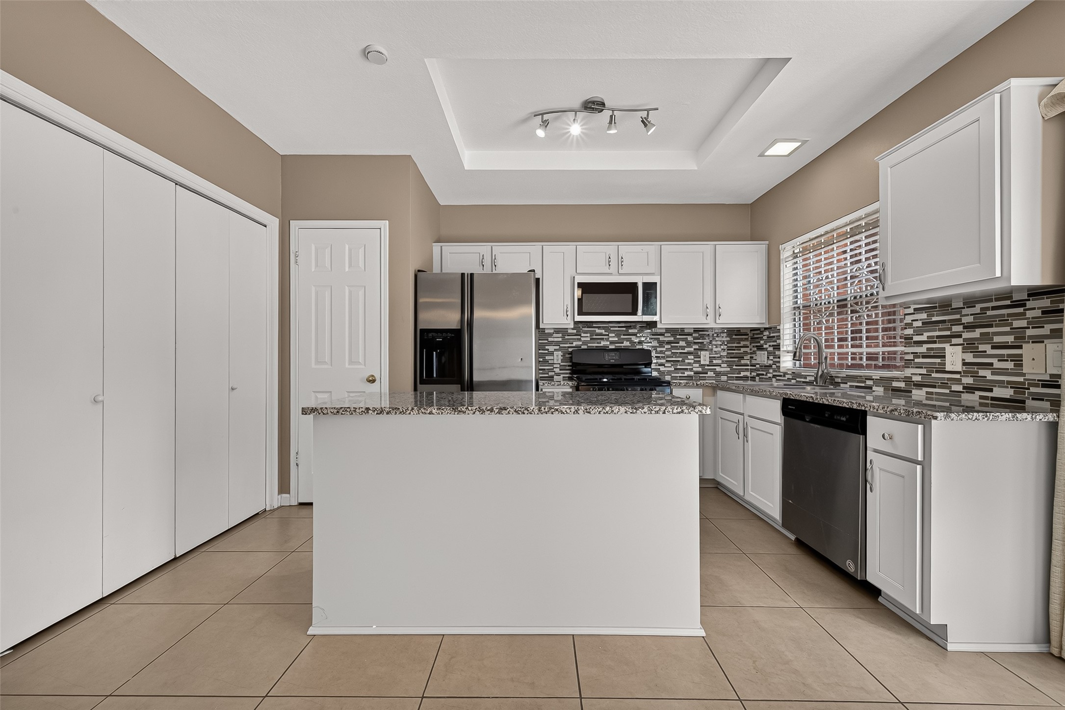 Spacious kitchen featuring granite countertops, center island, white cabinetry, tile backsplash, stainless-steel refrigerator and microwave, and ample cabinet and counter space.