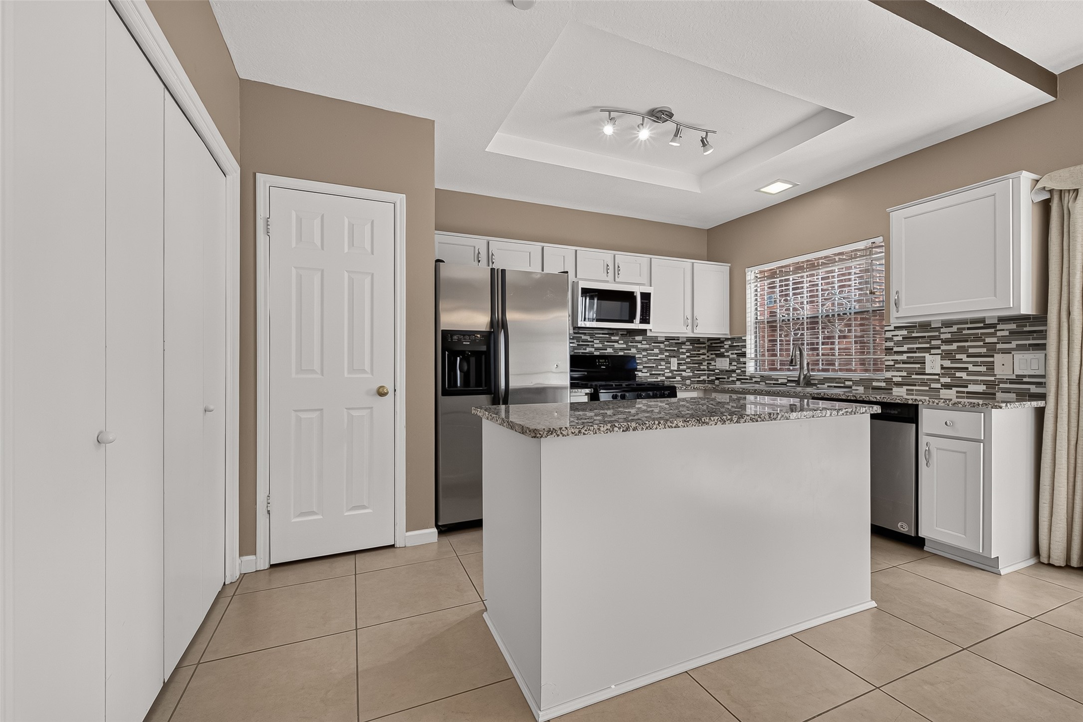 Another view highlighting the generous granite-topped center island, abundant cabinetry, stainless-steel appliances, decorative backsplash, and tray ceiling with contemporary lighting.