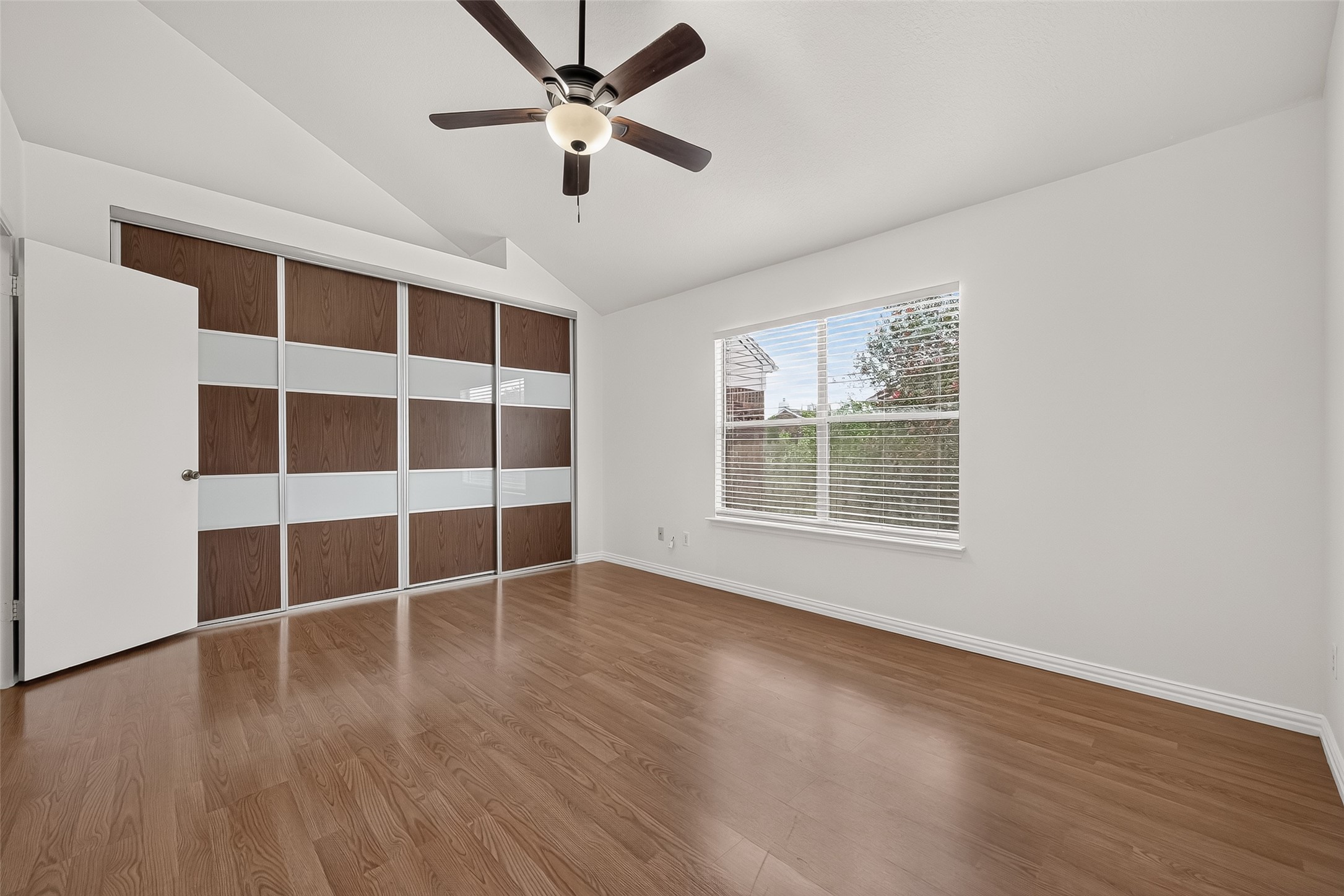 Bright primary bedroom with vaulted ceiling, large window, wood-look flooring, and impressive newly installed sliding closet doors providing a clean, contemporary look.