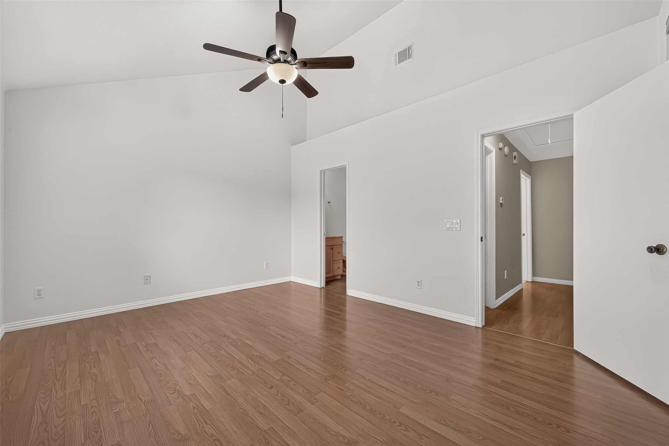 Generously sized primary suite with vaulted ceiling, ceiling fan, wood-look flooring, neutral finishes, and convenient access to the en-suite bath.