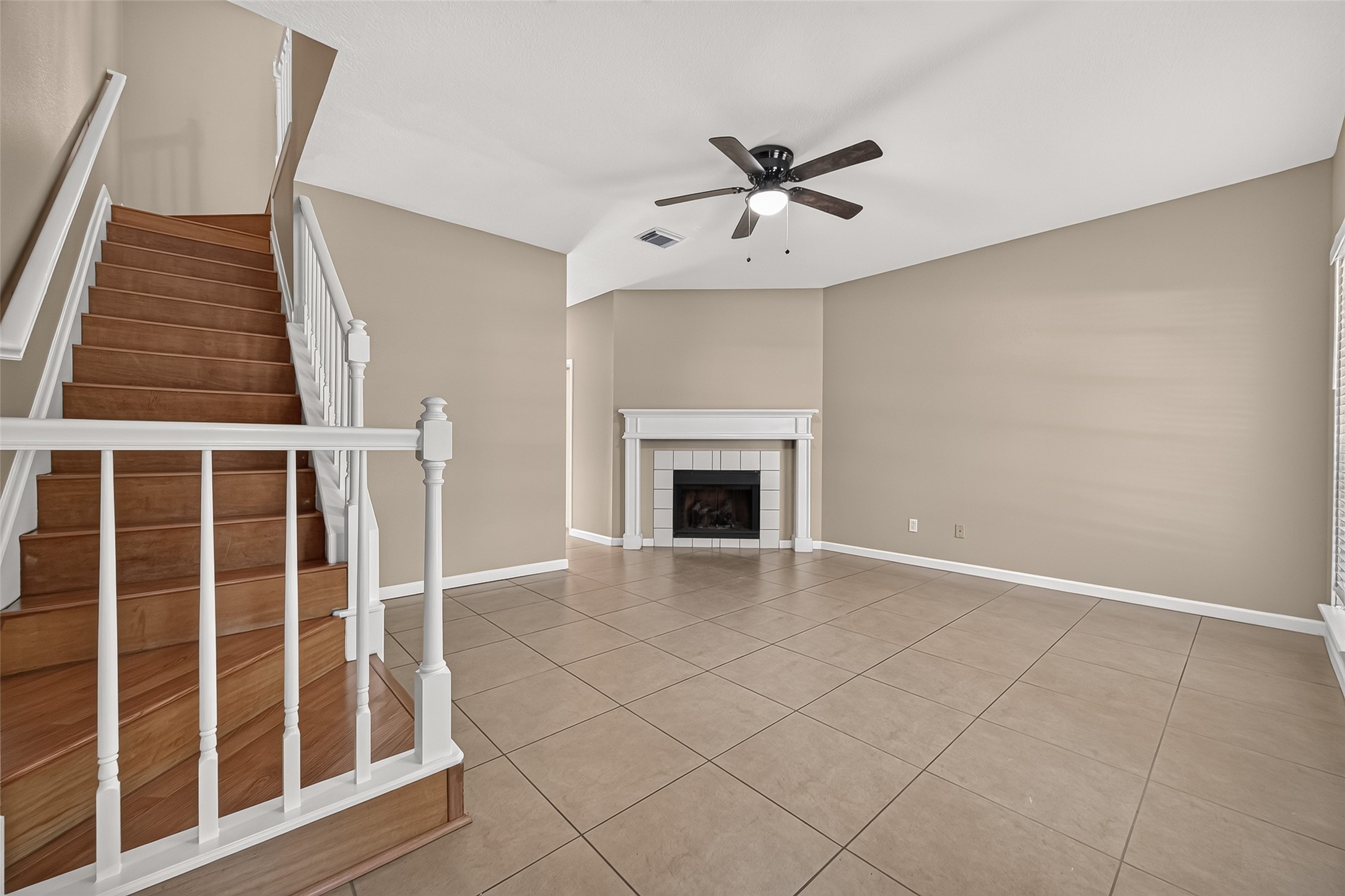 Open living area featuring tile flooring, fireplace, ceiling fan, neutral finishes, and attractive wood staircase with white railing leading to the second floor. Open living area featuring tile flooring, fireplace, ceiling fan, neutral finishes, and attractive wood staircase with white railing leading to the second floor.