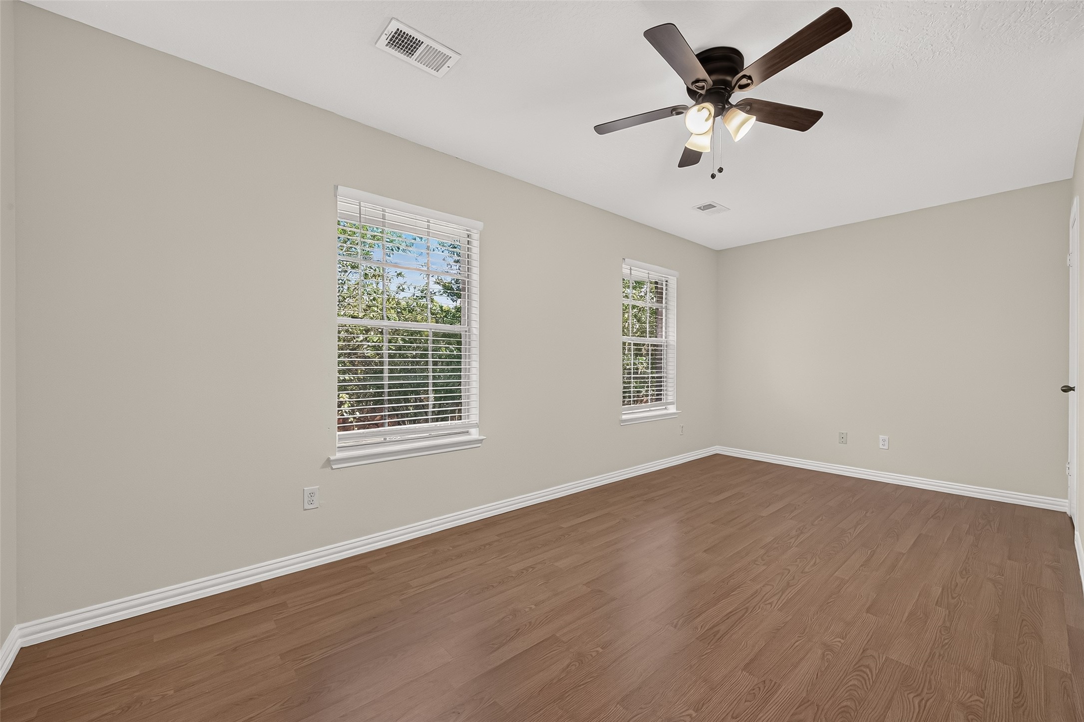 Bright and generously sized secondary bedroom featuring wood-look flooring, ceiling fan, neutral finishes, and multiple windows providing abundant natural light.