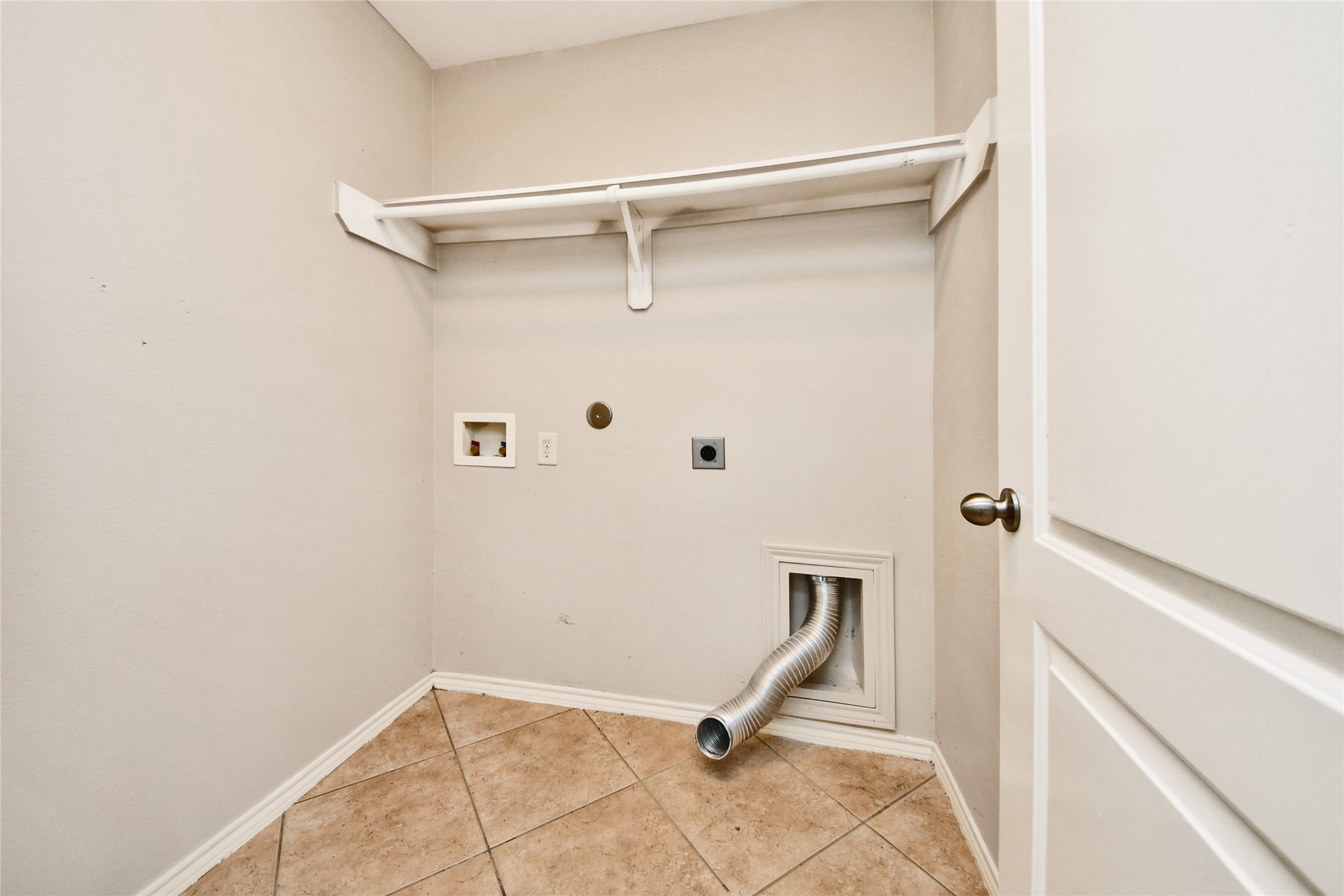 Spacious laundry room with tile flooring, ample storage shelf, and hookups for washer and dryer.