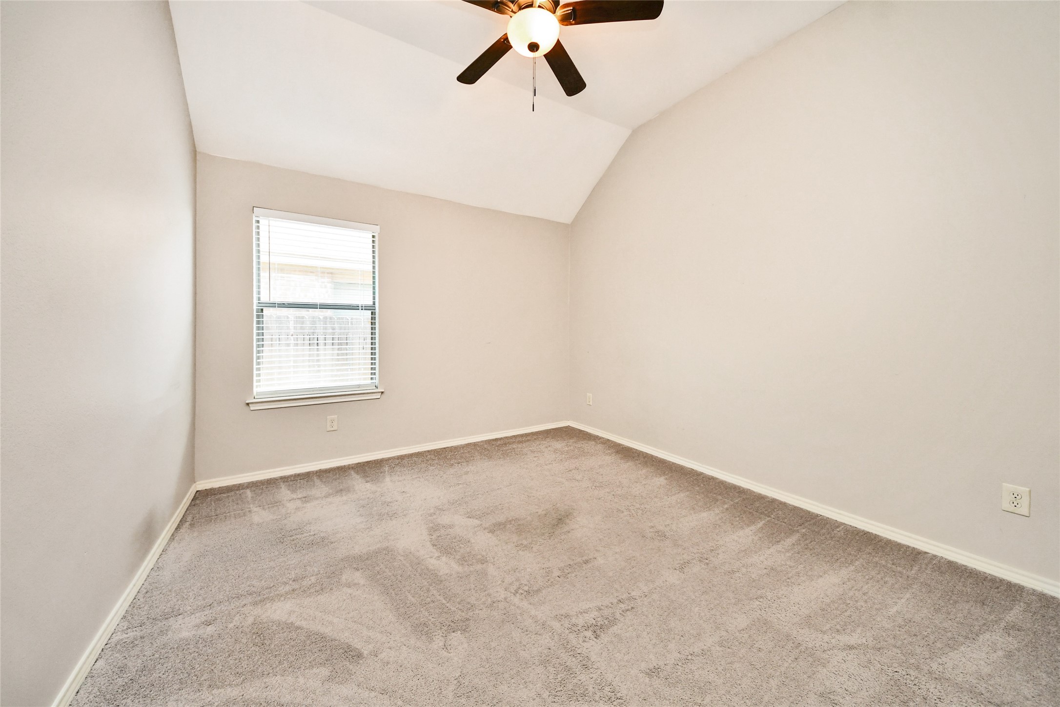 Spacious bedroom with plush carpeting, vaulted ceiling, and natural light from a single window, enhanced by a modern ceiling fan.
