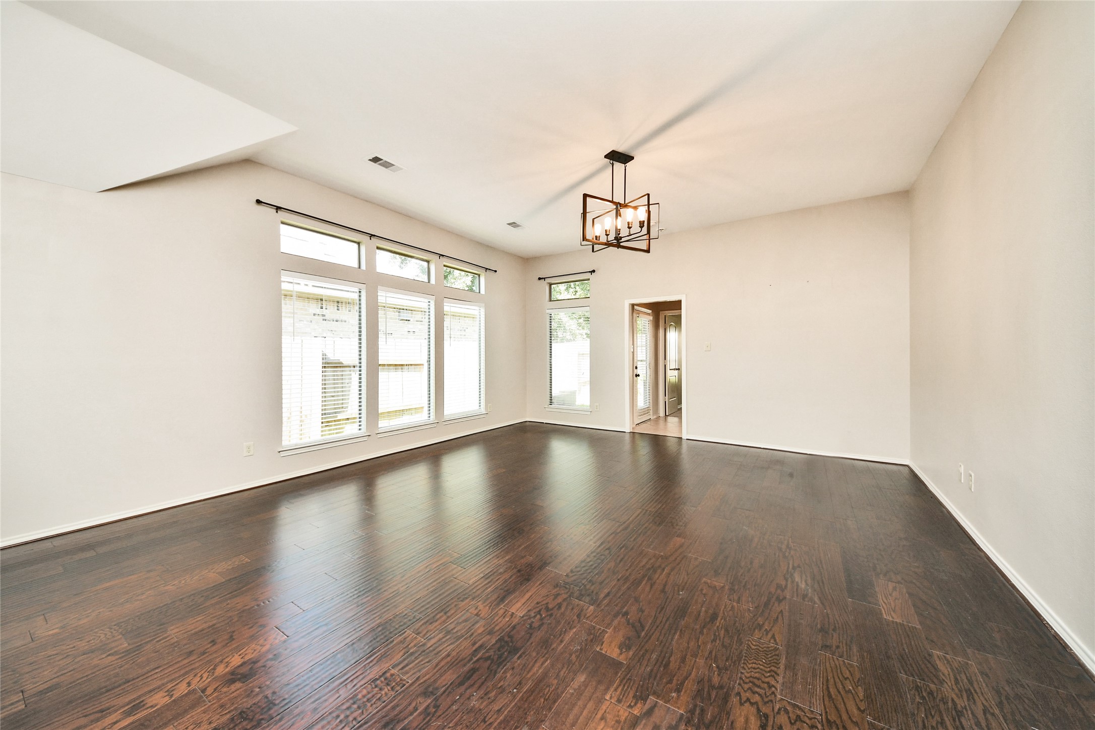 Spacious living area with rich hardwood floors, abundant natural light from large windows, and a modern light fixture, perfect for comfortable gatherings.