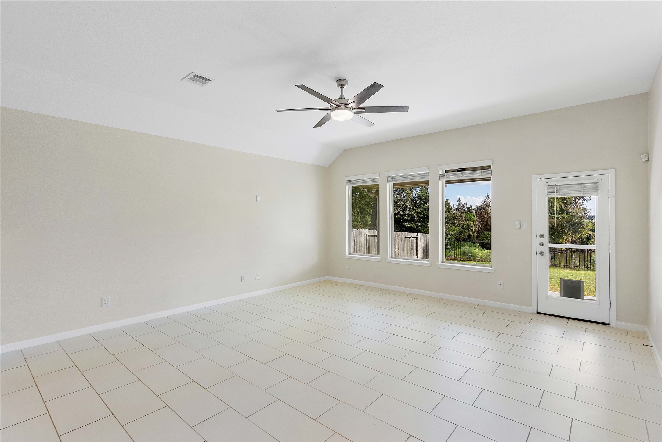 Spacious and bright living area with large windows, modern ceiling fan, and tile flooring, offering direct access to a lush backyard.