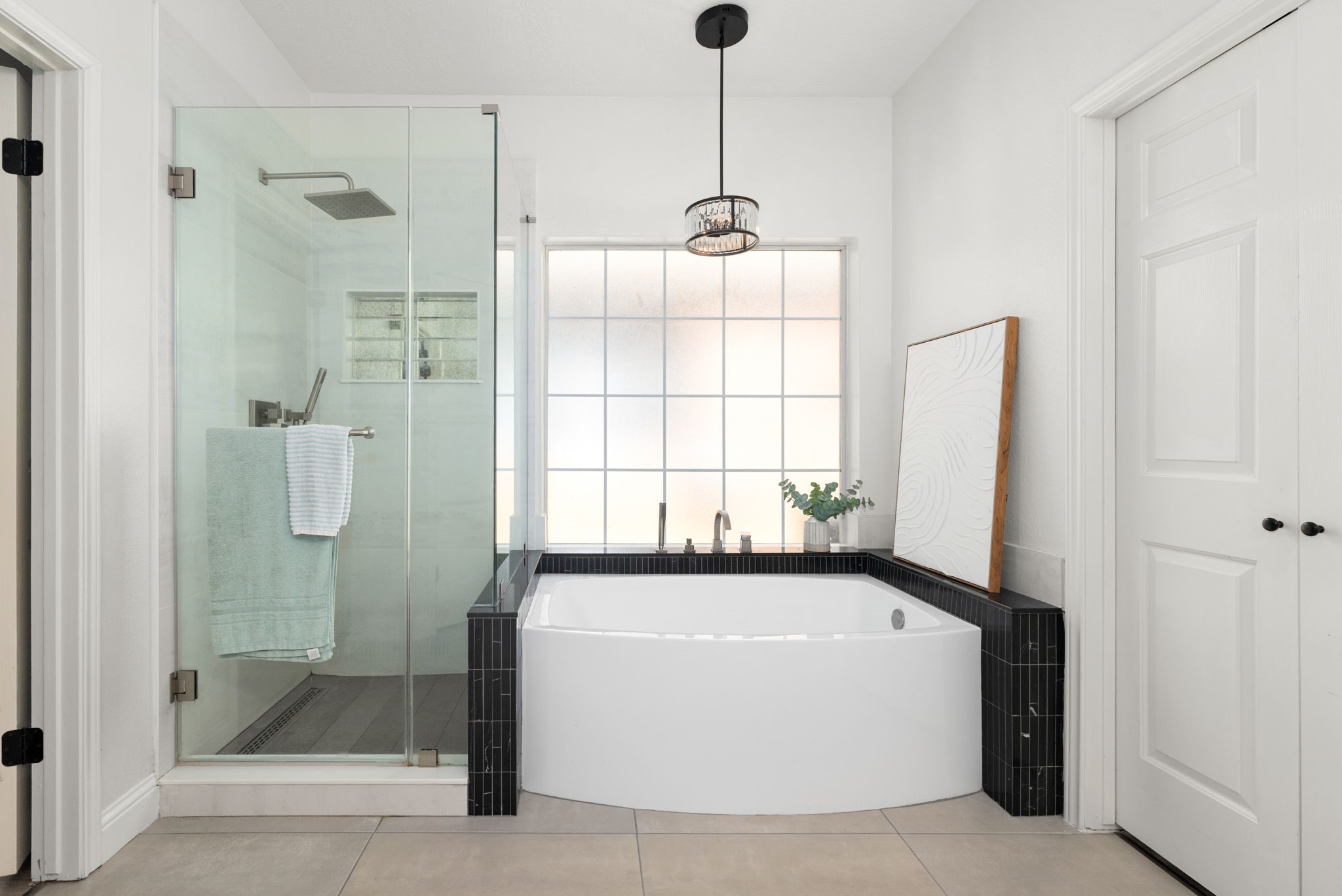 This modern bathroom features a luxurious soaking tub framed by sleek black tiles and a spacious glass-enclosed shower, complemented by a large frosted window for natural light and privacy.