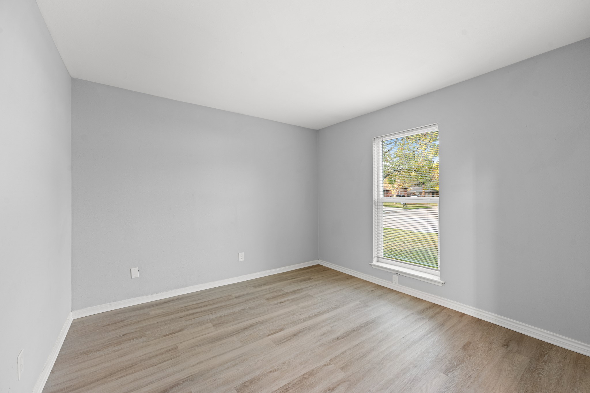 A bright, versatile home office features wood-look flooring, neutral gray walls, and a large window overlooking the front yard, creating an inviting space for working from home.