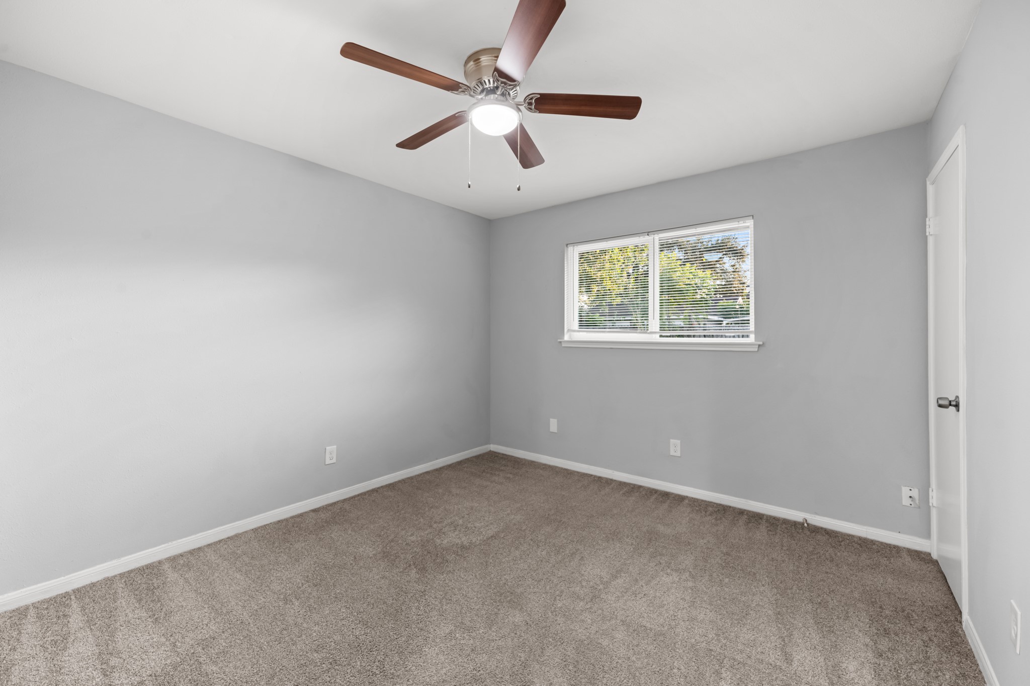 This comfortable additional bedroom offers plush carpeting, neutral gray walls, a ceiling fan with integrated lighting, and a window overlooking the outdoors.