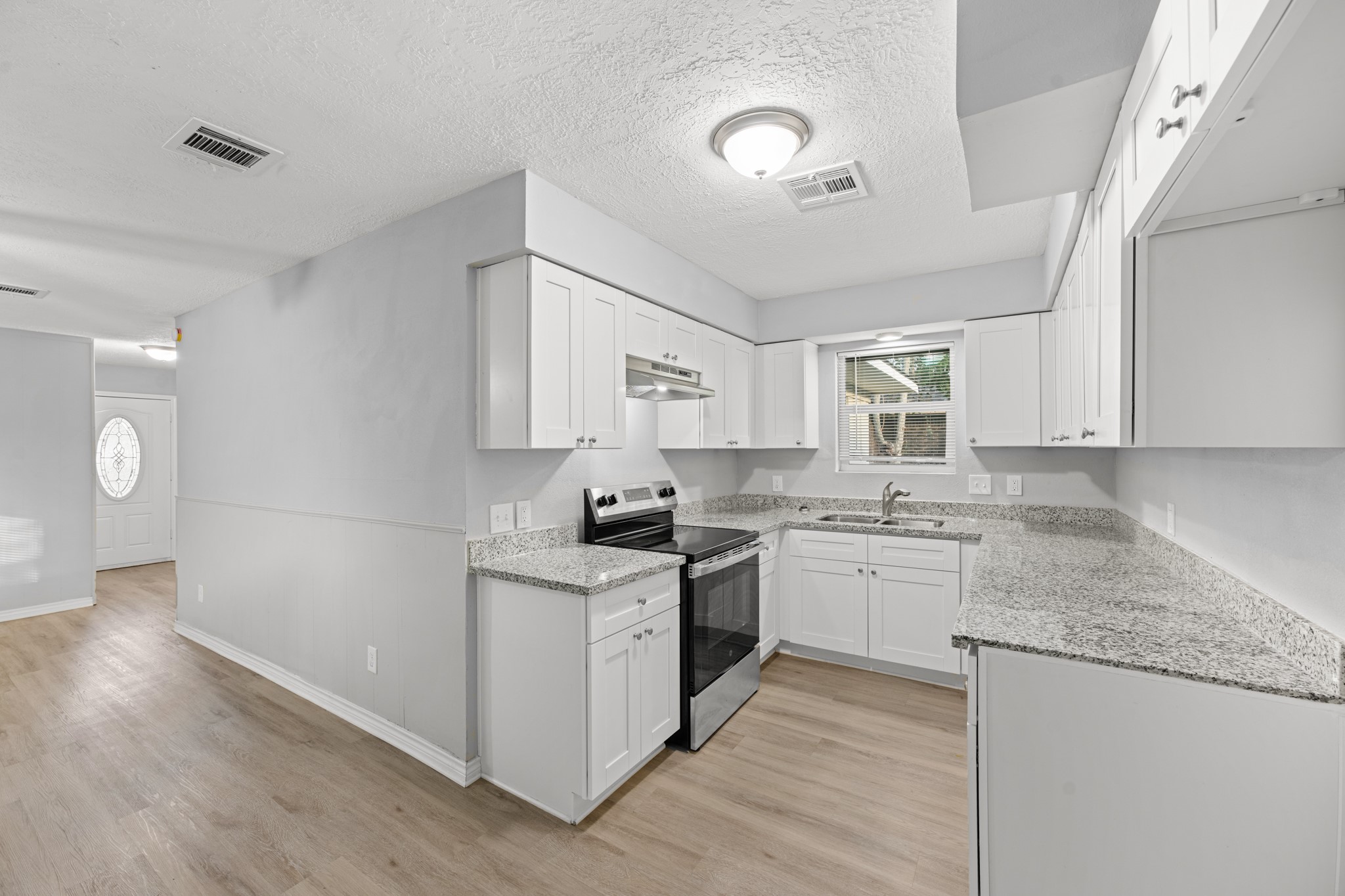 The beautifully updated kitchen features white shaker-style cabinetry, granite countertops, stainless steel appliances, and wood-look flooring, with an open connection to the adjoining living area.