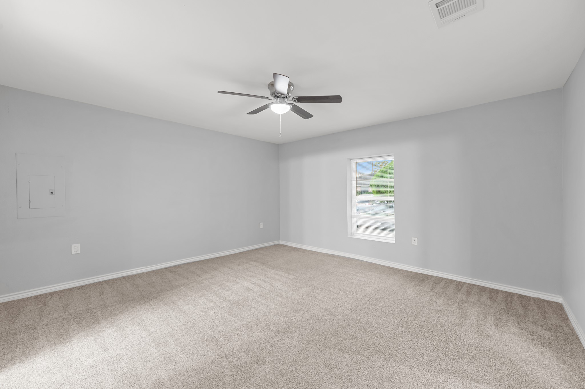 The spacious primary bedroom offers plush neutral carpeting, soft gray walls, a ceiling fan with lighting, and a window that welcomes natural light into the room.