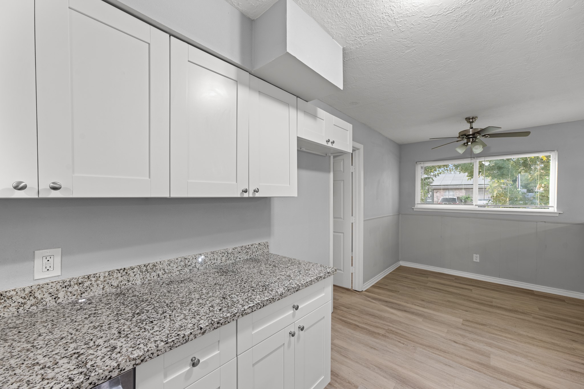 The kitchen's extended granite countertop provides additional workspace and storage, while the adjoining dining area features a large window, ceiling fan, and classic wainscoting.